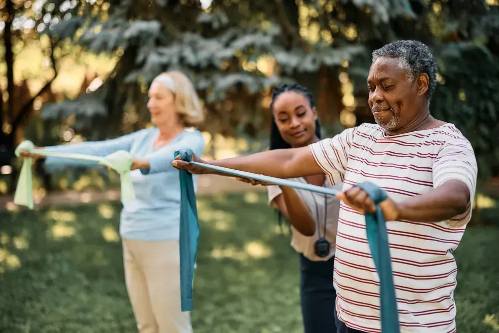 An older Black man and a white woman doing exercises with resistance bands, supervised by a young Black woman in a park.