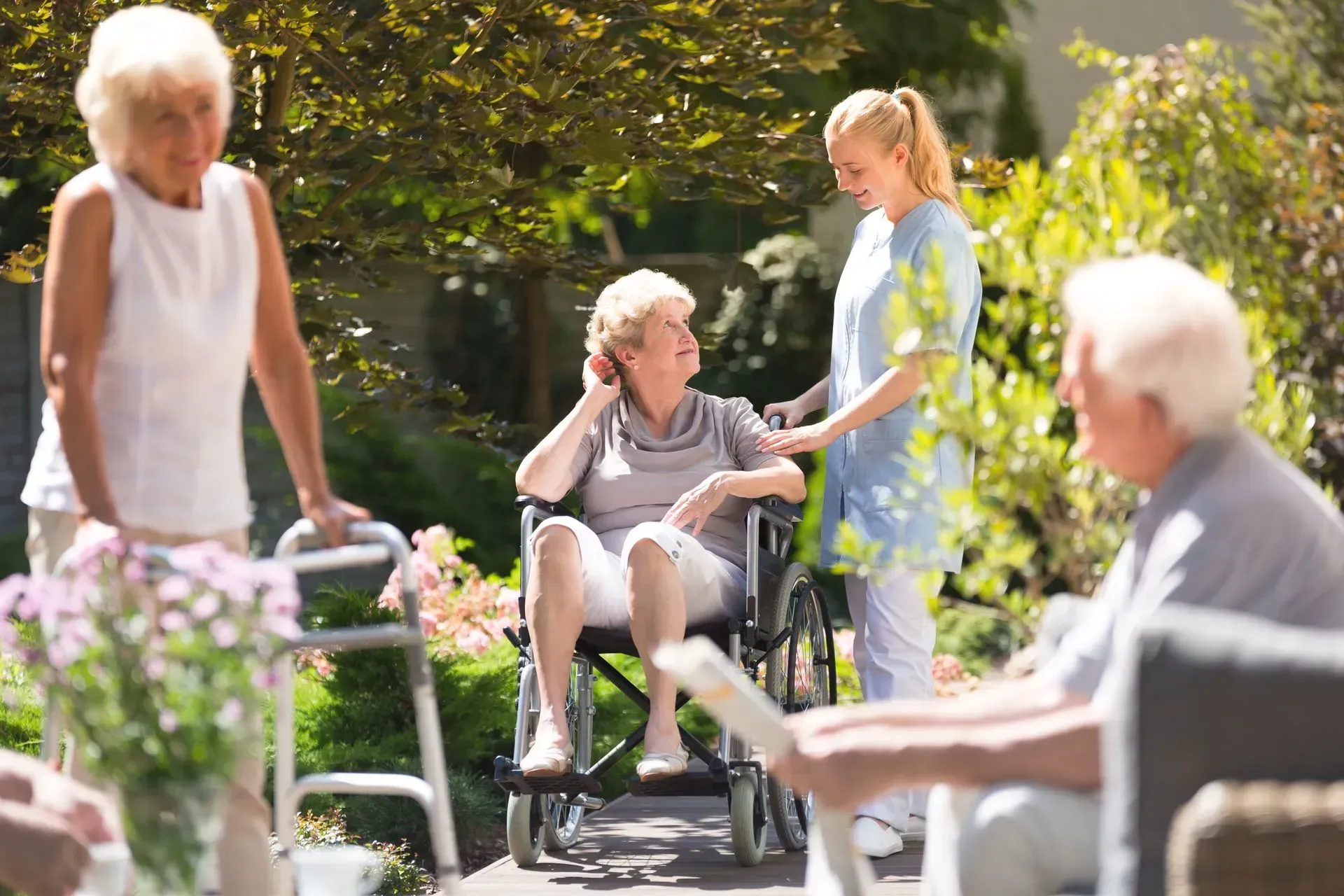 Seniors with caregiver outdoors: woman in wheelchair, others with walker and reading. Sunny garden setting.