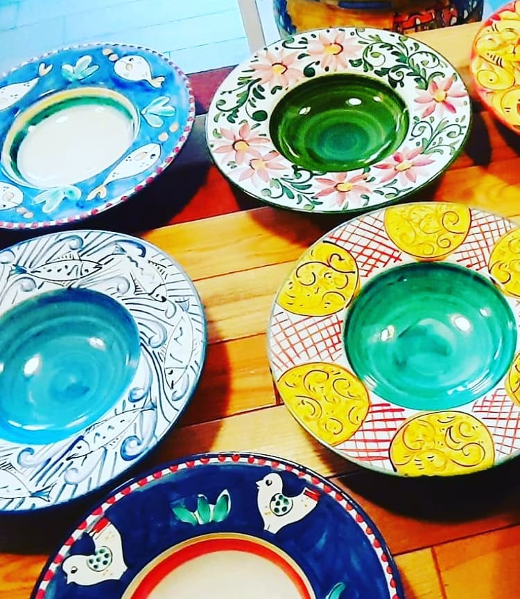 Ciotole colorate in ceramica dipinta a mano su una superficie di legno. I motivi includono fiori, uccelli e motivi geometrici.
