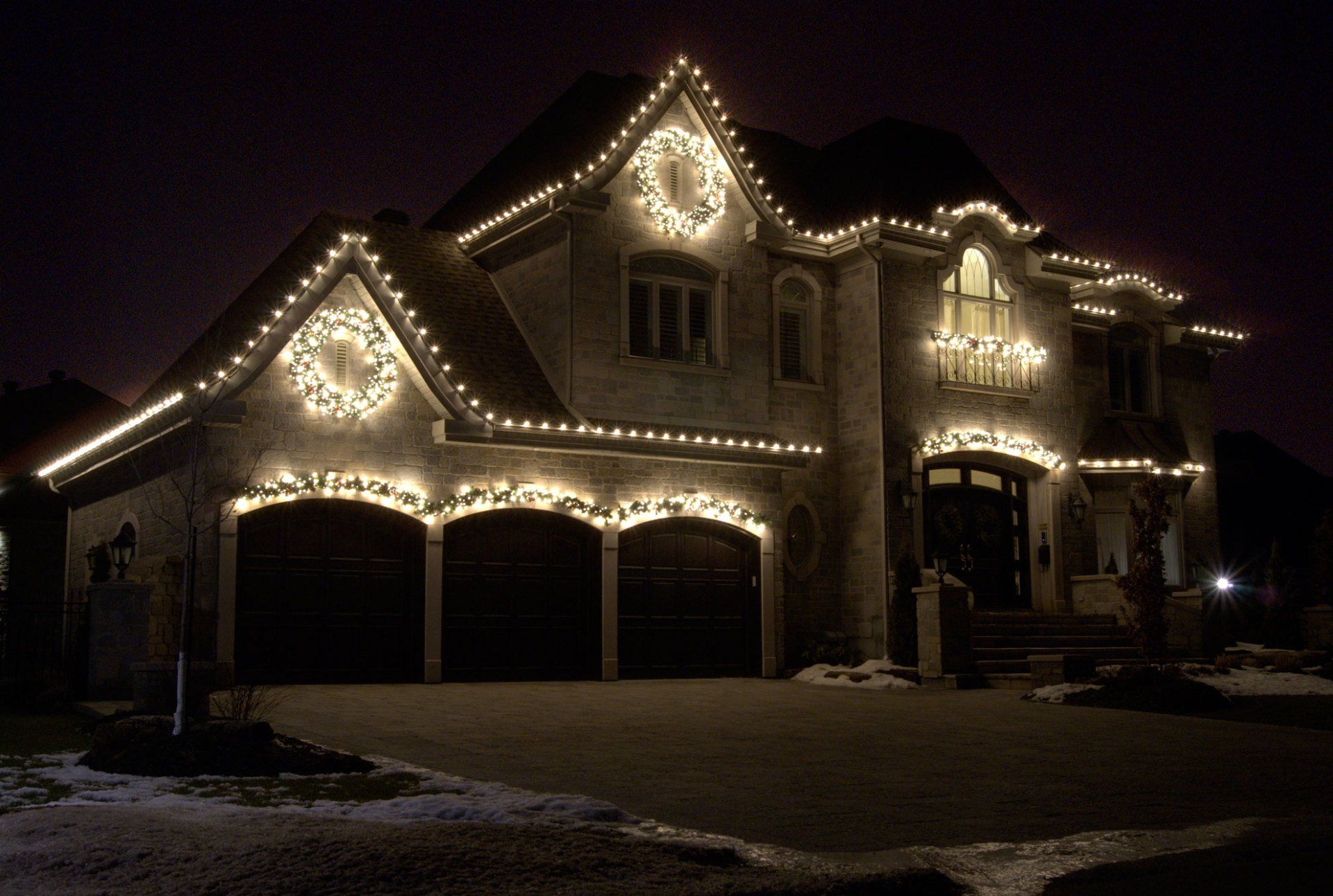 House decorated with Christmas lights, featuring wreaths and string lights along the roof and garage.