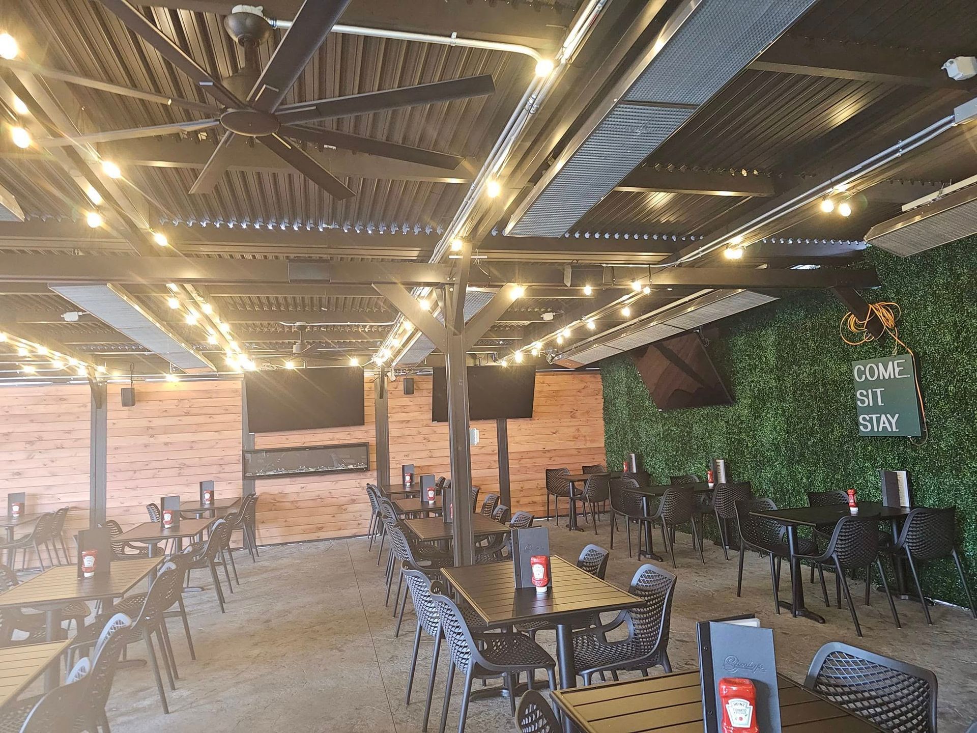 Outdoor dining area with tables, chairs, TVs, and string lights under a covered patio.