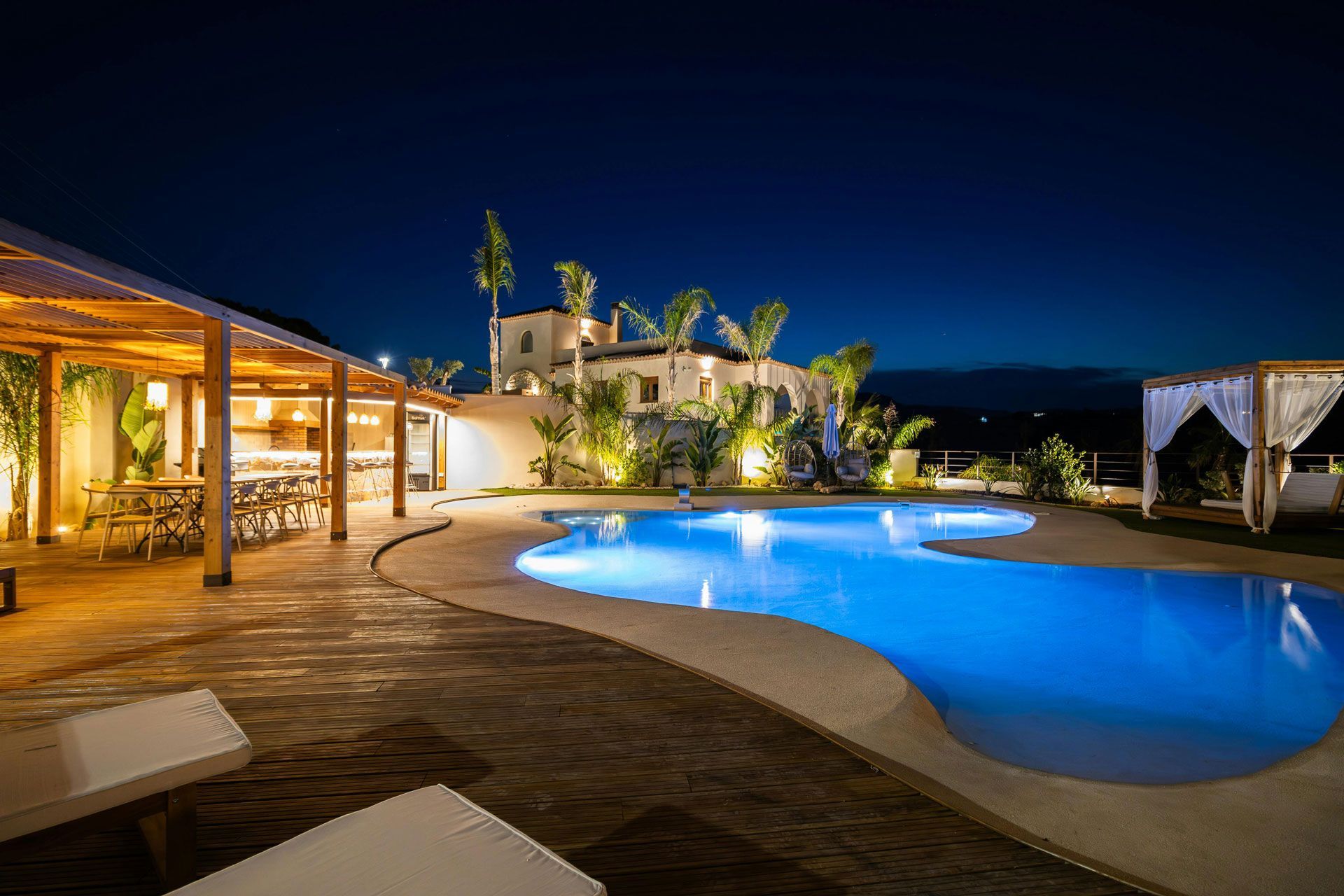 Night view of a resort pool, white building, patio seating, and a pergola with lights against a dark blue sky.