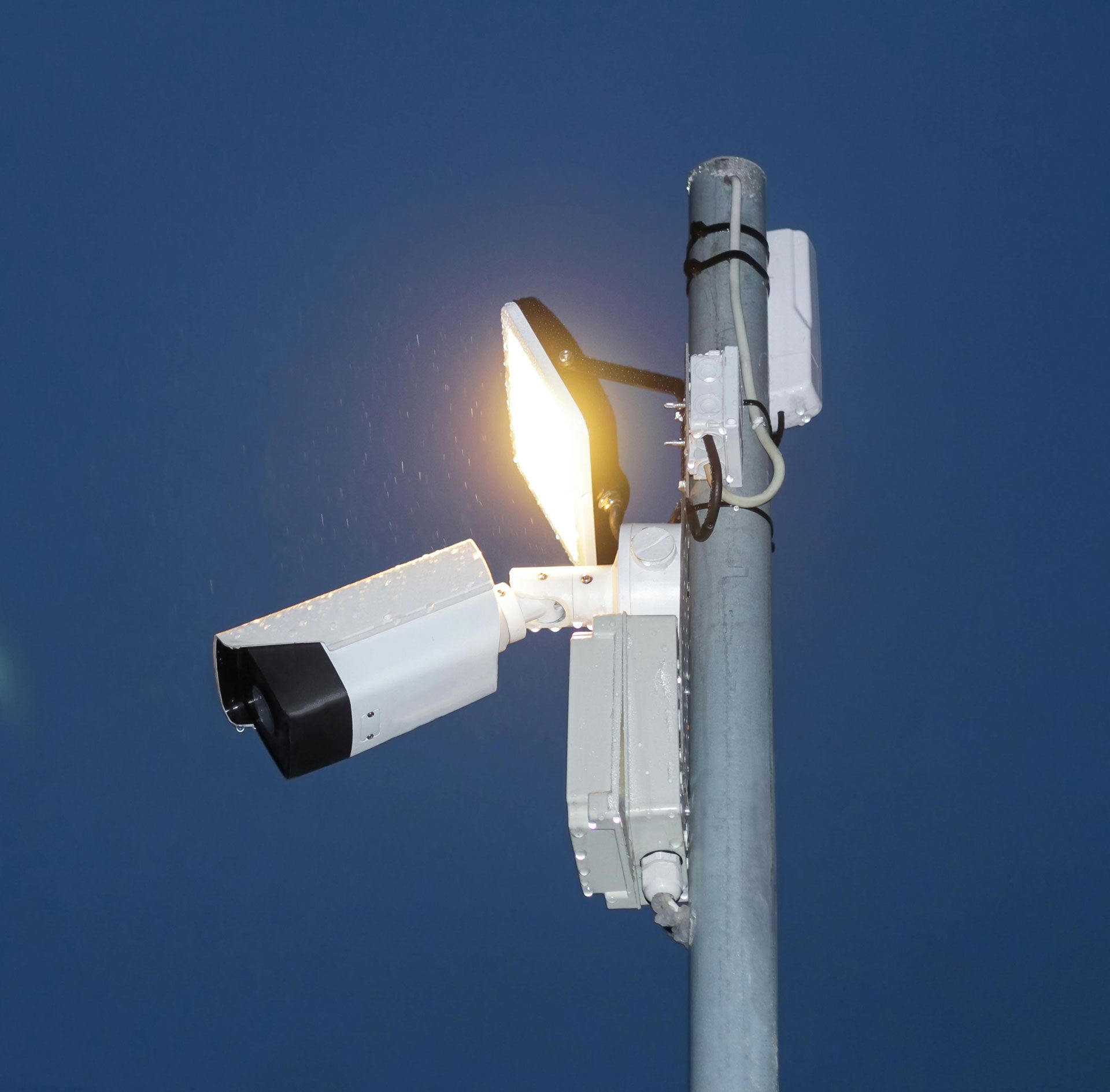 Security camera and spotlight mounted on a pole against a dark blue sky.