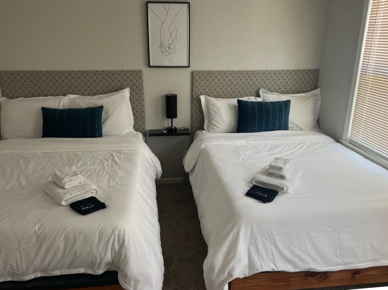 Two neatly made twin beds in a bright hotel room with blue pillows and a nightstand between them.