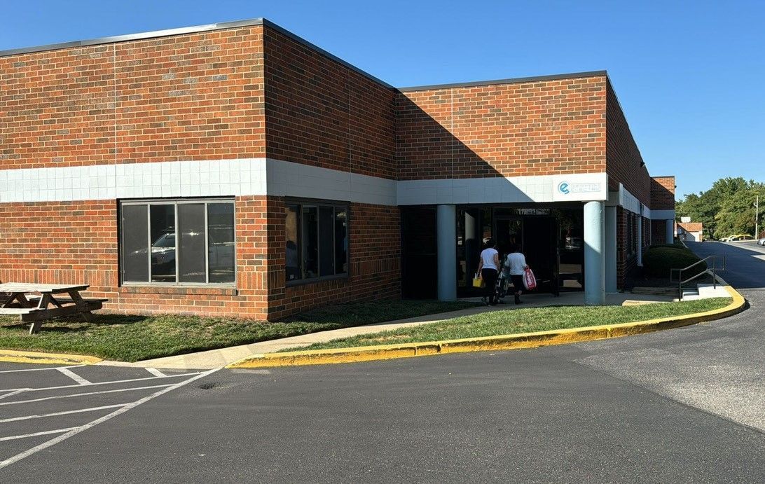 Brick building with white trim and covered entrance beside a parking lot on a sunny day