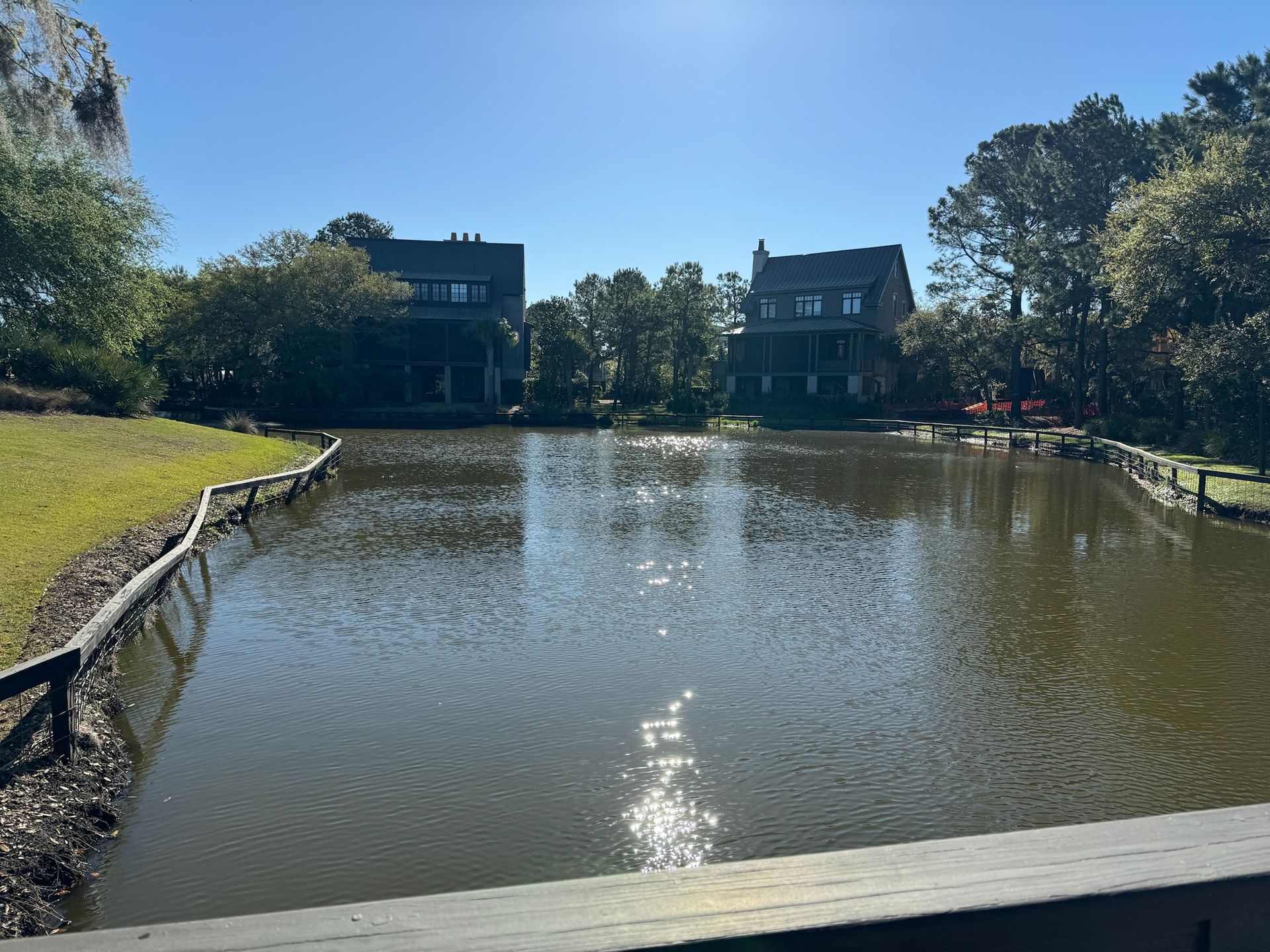 Two-story houses overlook a calm pond on a sunny day. A wooden fence lines the pond's edges.