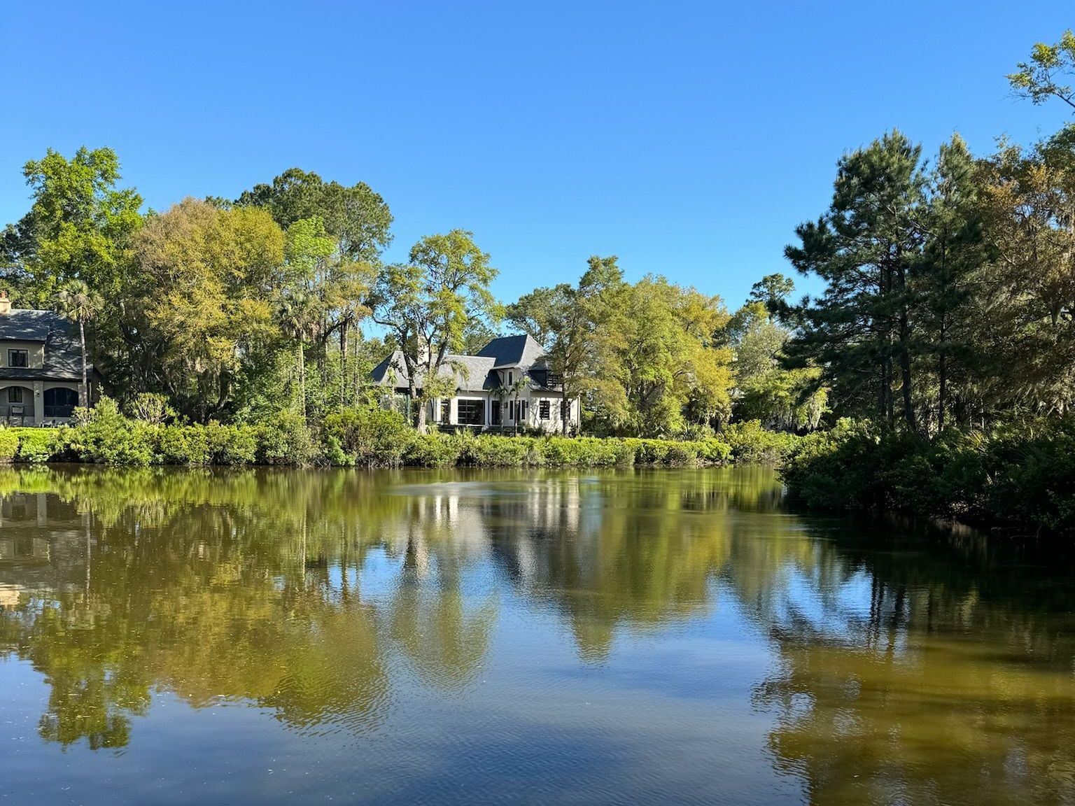 A calm lake reflects a house and surrounding trees under a clear, blue sky.