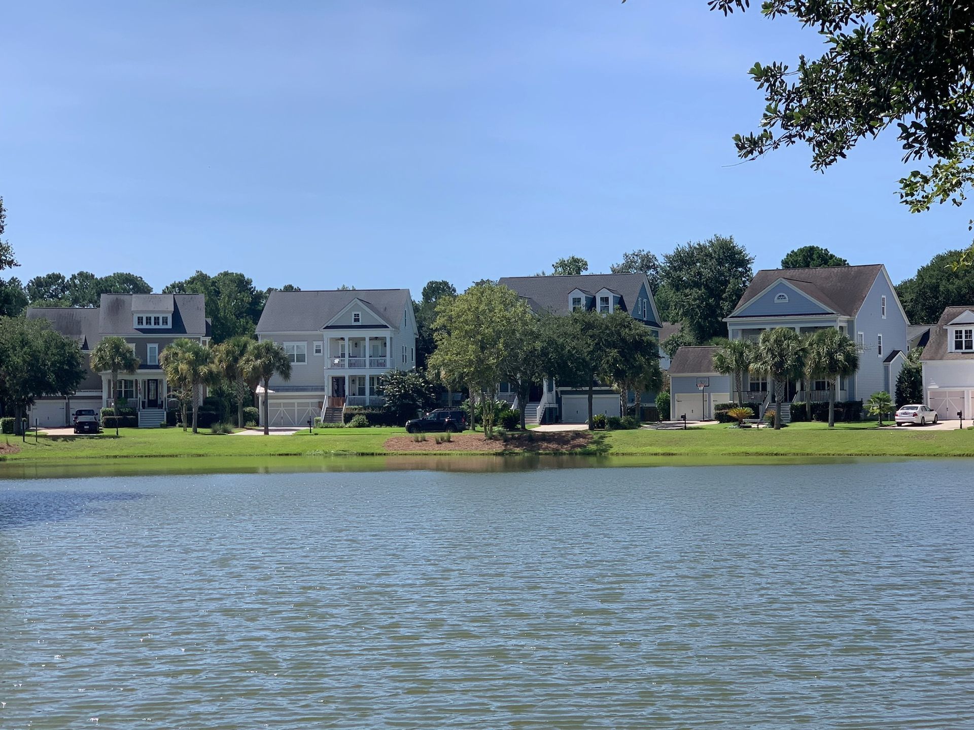 Houses with light-colored siding line a lake under a clear, blue sky, some with garages.