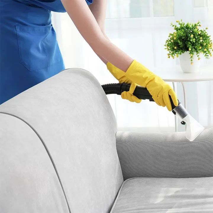 Yellow-gloved cleaner vacuuming a gray couch in a bright living room