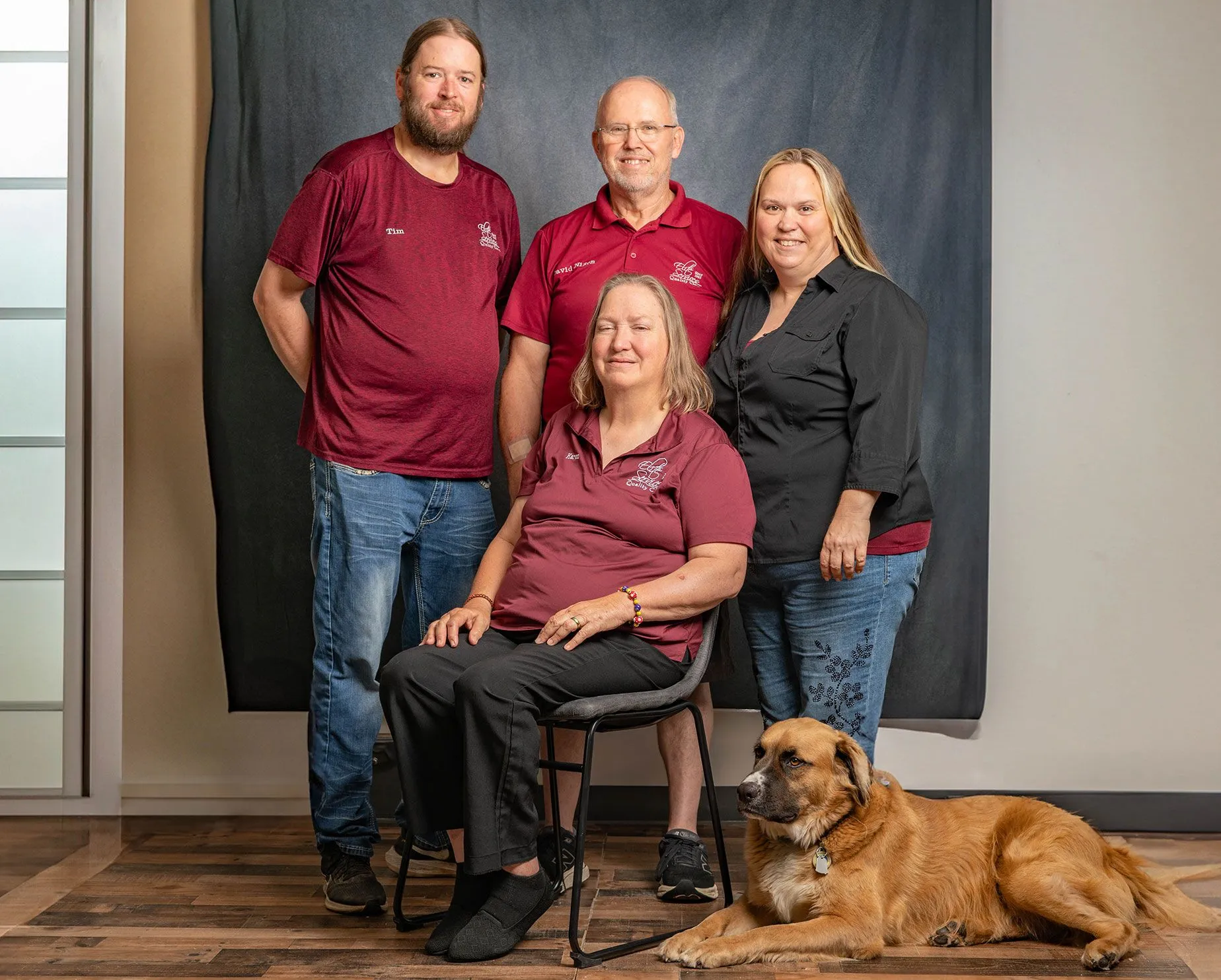 Four people pose in front of a dark backdrop, with a brown dog lying on the floor in front.