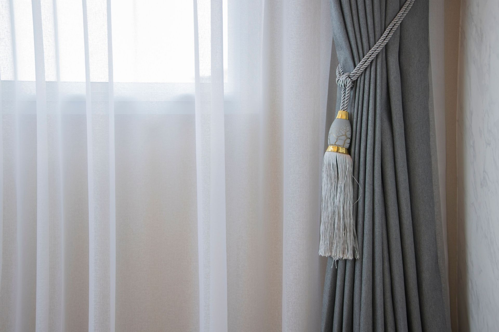 Sheer white curtains beside gray drapes with a decorative tassel tieback by a window