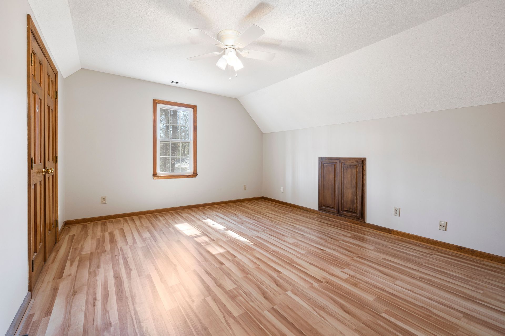 Empty carpeted room with sloped white ceiling, hardwood floor, window, and wooden closet doors