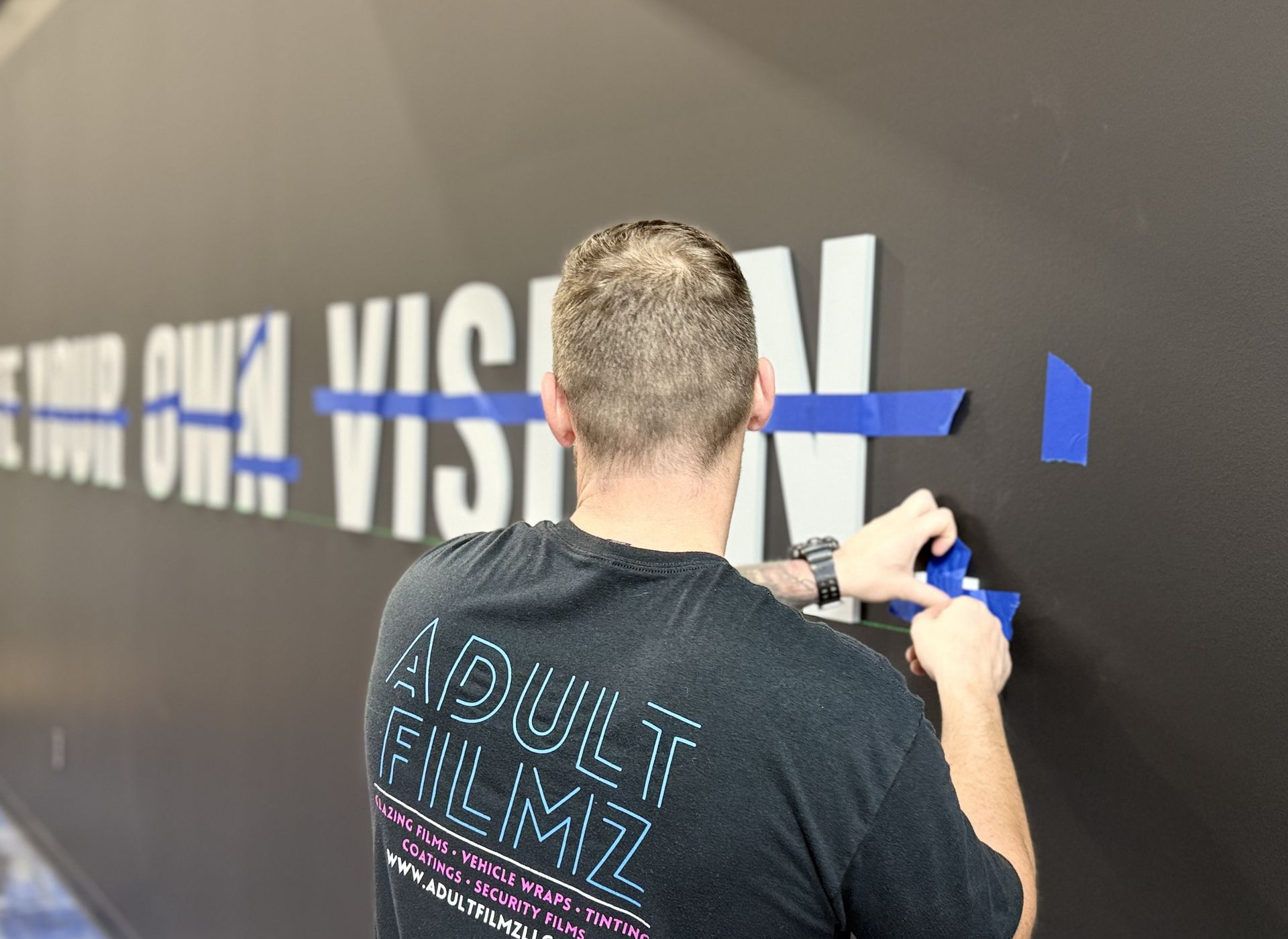 Person applying blue tape to black wall lettering during a mural installation