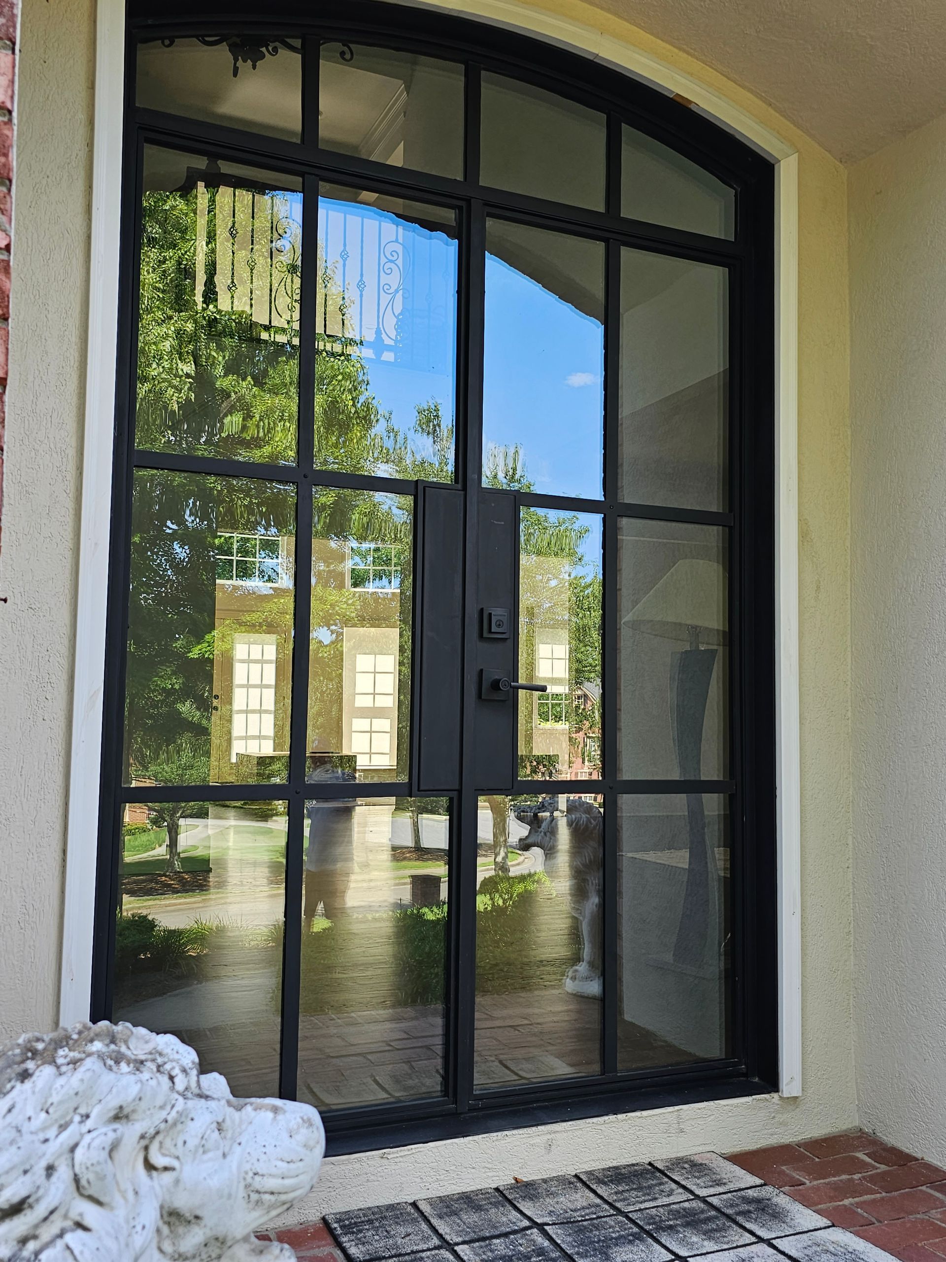 Black arched glass front door reflecting a courtyard and greenery, with a stone lion statue nearby