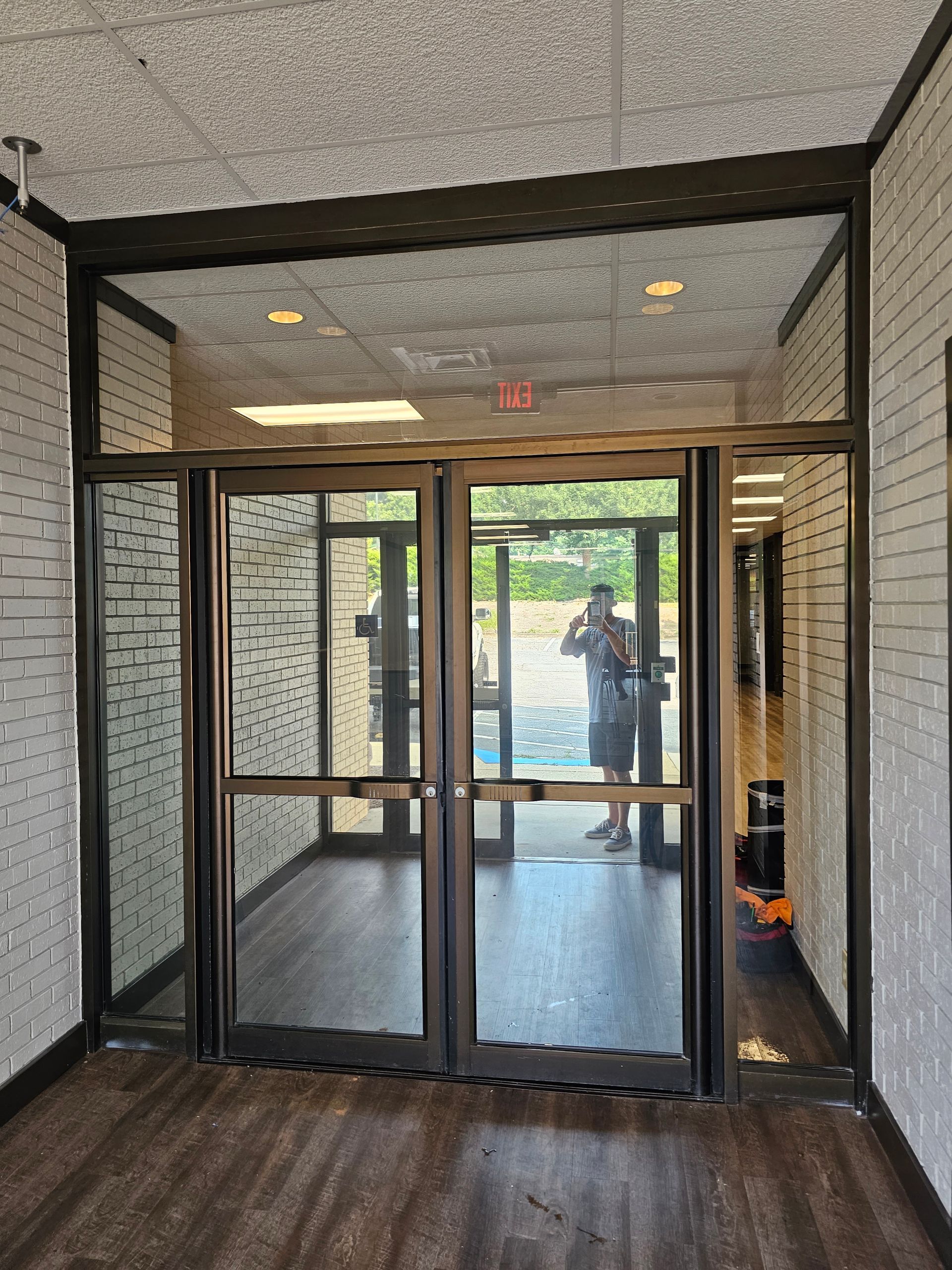 Glass lobby entrance with black framing, tiled ceiling, wood floor, and a person reflected in the doors