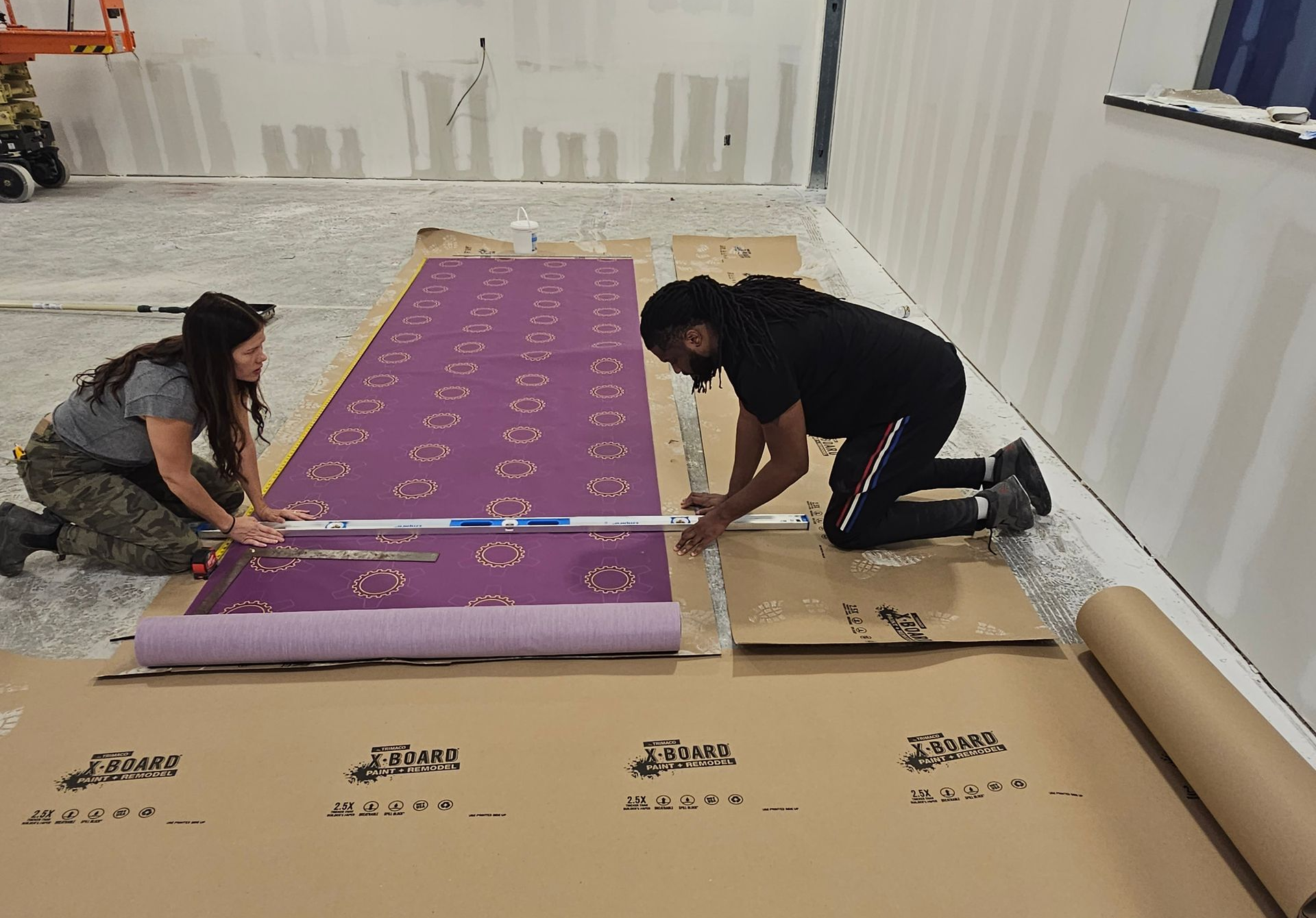 Two people unrolling a purple carpet in a white room with protective floor covering