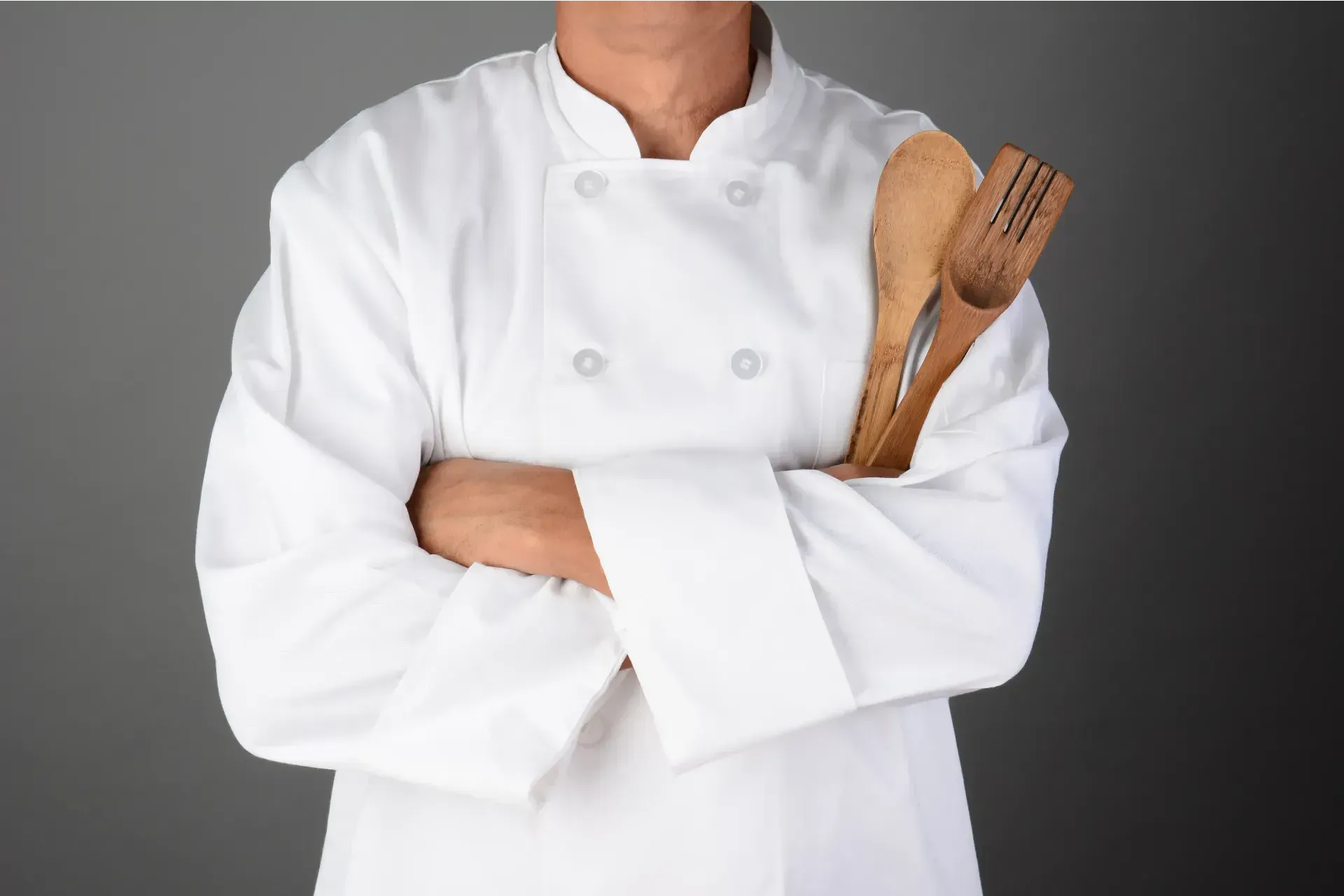 Chef in white uniform, arms crossed, holding wooden spoon and fork, gray background.