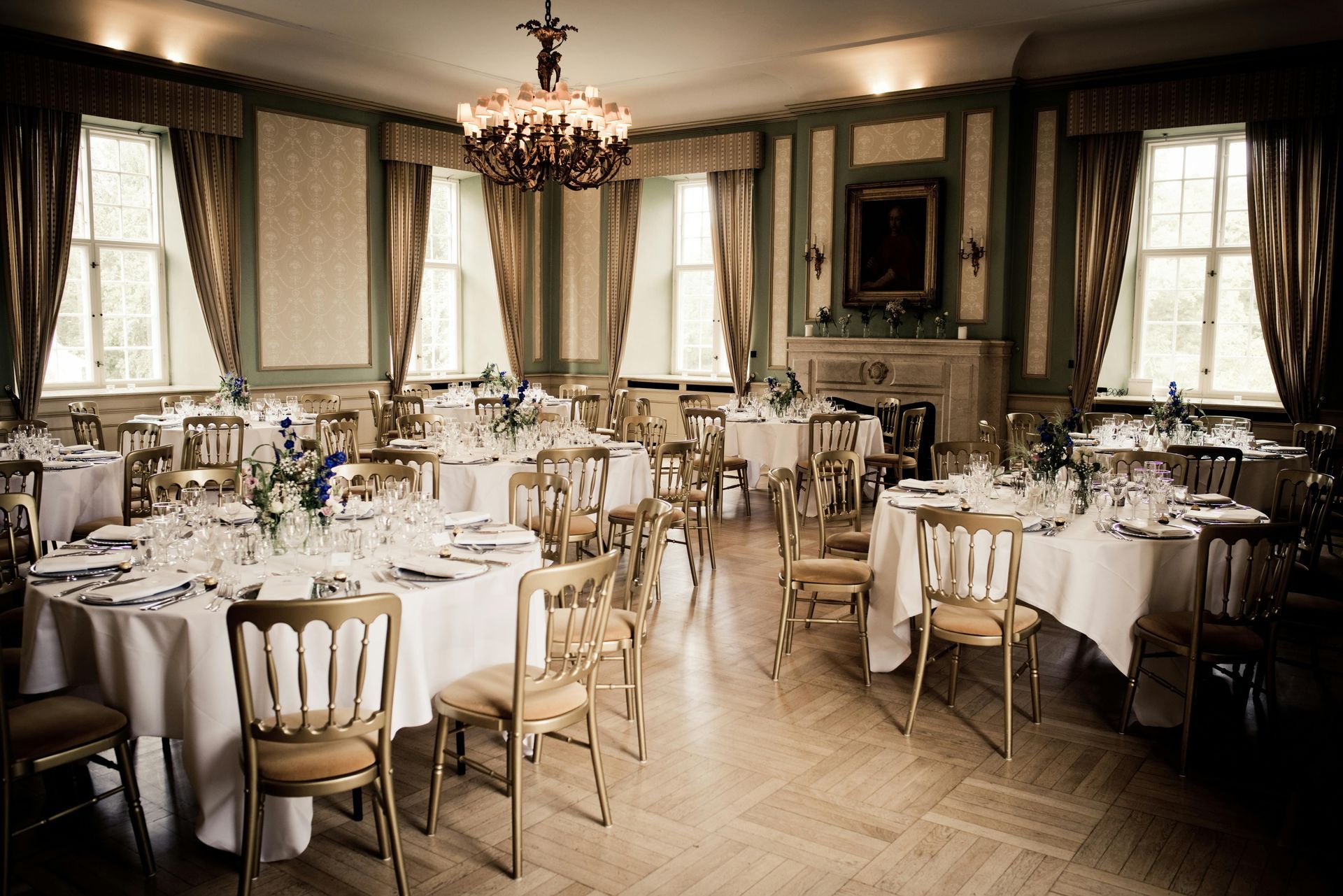 Elegant dining room set for a celebration. Round tables with white linens, gold chairs, and floral centerpieces.