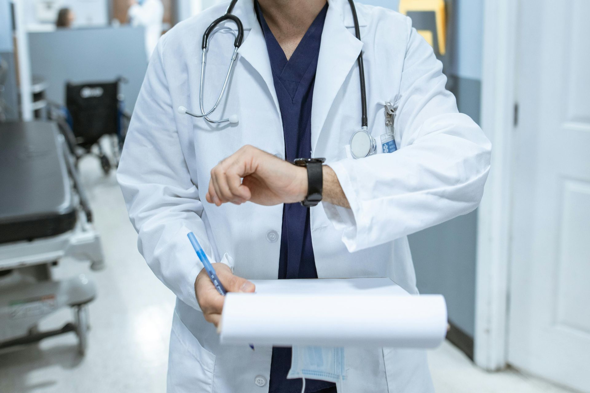 Doctor in white coat looking at watch, holding clipboard in a hallway with medical equipment.