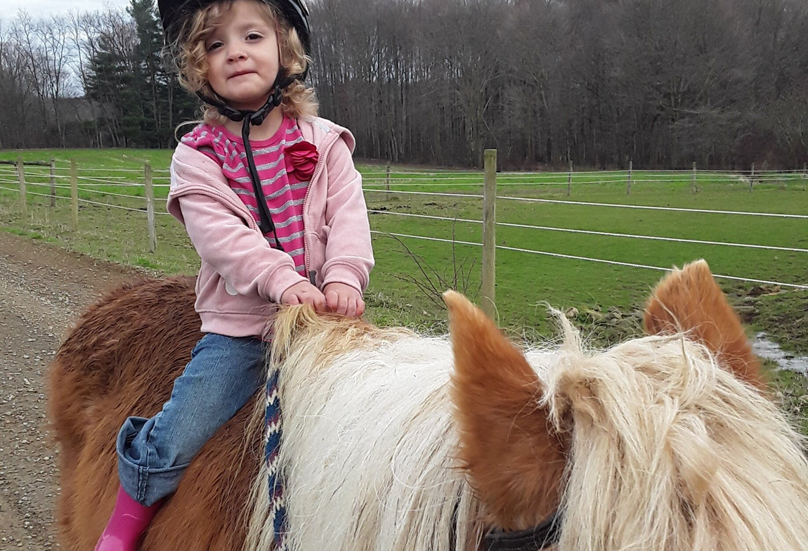 Child riding a brown-and-white pony in a grassy field, wearing a pink jacket and smiling.