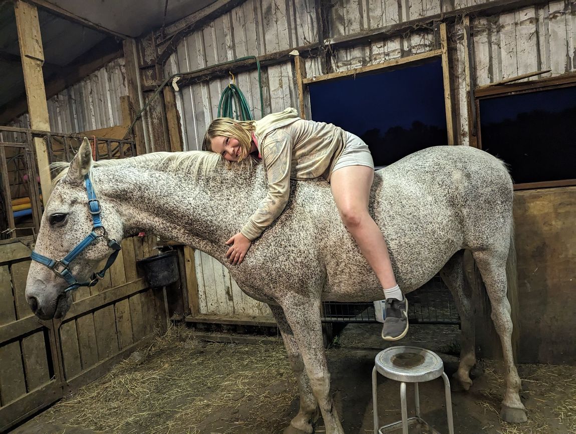 Person lying on a gray horse in a barn, smiling while holding its neck.
