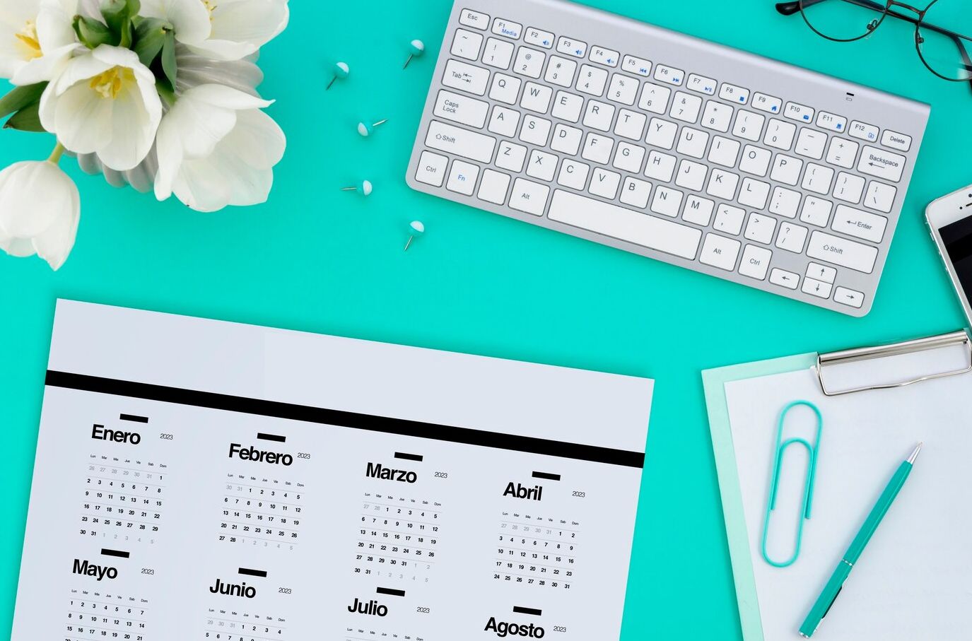 Un escritorio con calendario, teclado, celular, gafas y flores.