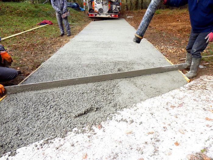Workers pouring and leveling wet concrete for a pathway, with a concrete truck in the background.