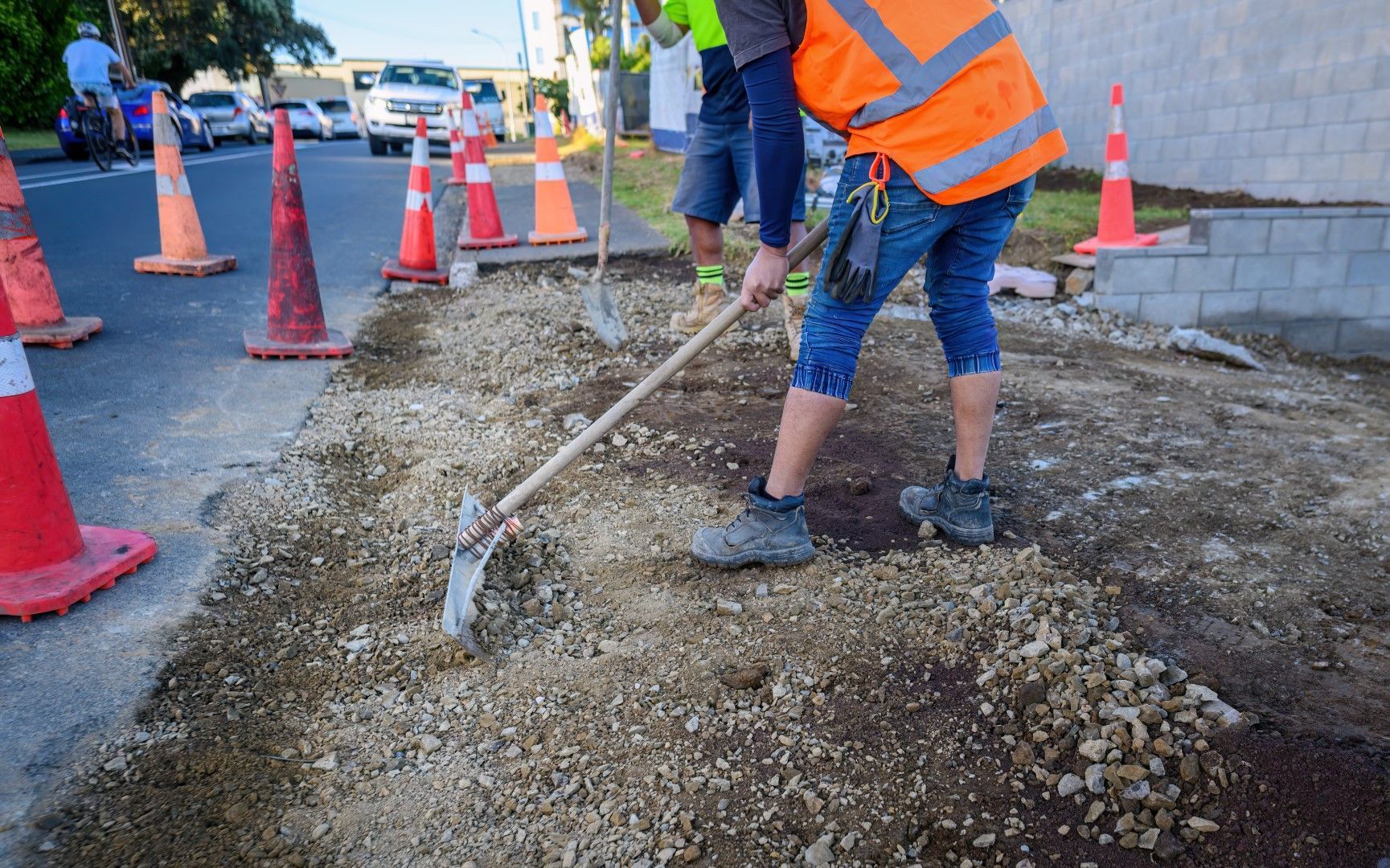Construction workers leveling gravel with shovels on a street next to orange traffic cones.