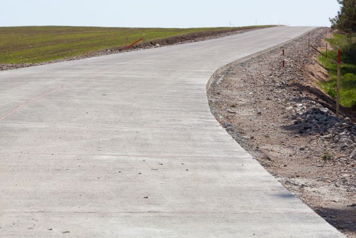 New concrete road curving uphill, with gravel shoulder and grassy bank. Sunny day.