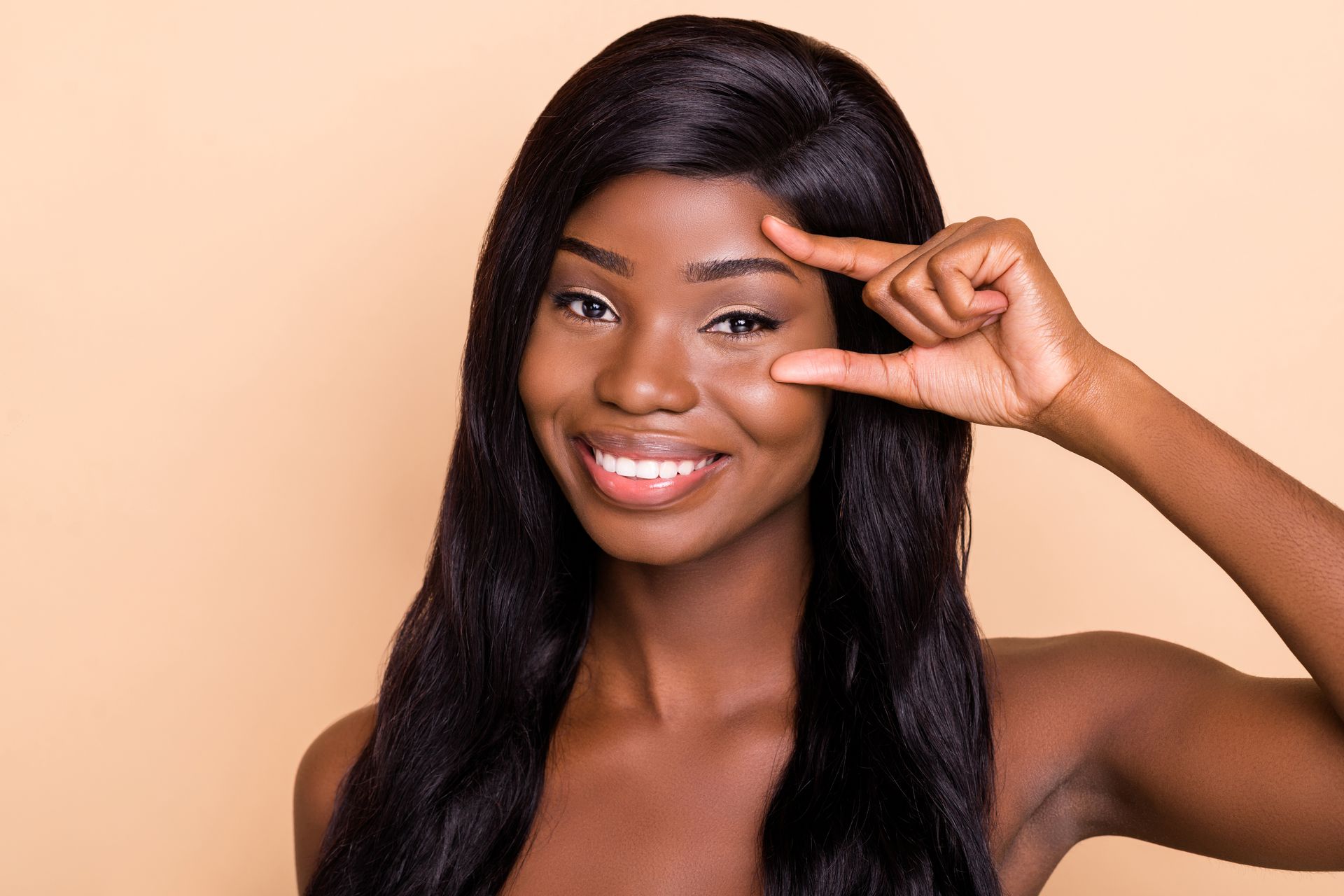 A woman with long black hair is smiling and holding her hand to her eye.