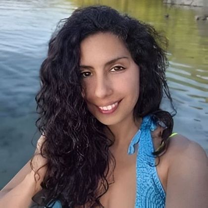 Persona sonriente de cabello oscuro y rizado, vestida de azul junto a aguas tranquilas