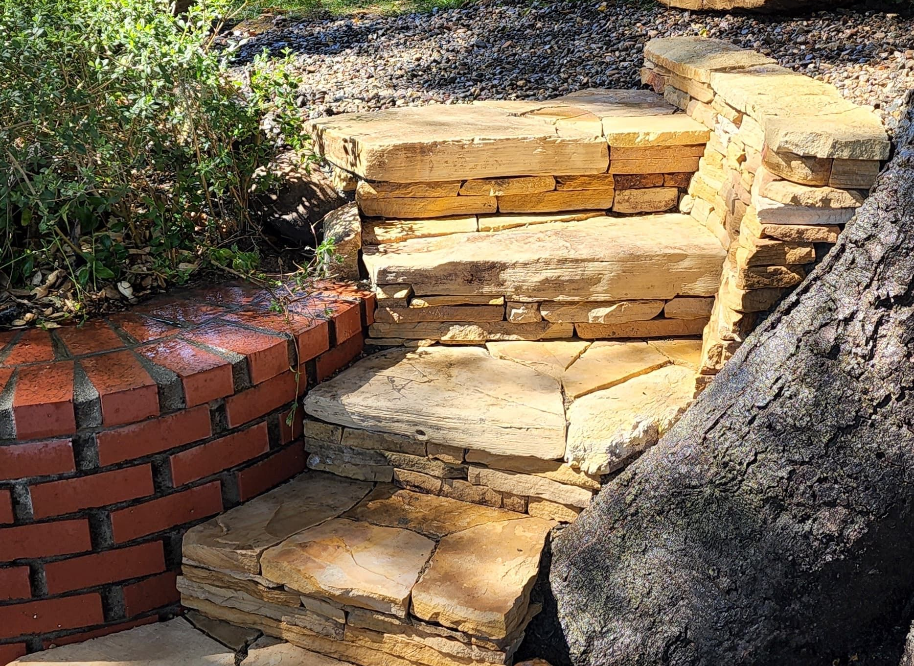 Stone steps integrated into a garden landscape next to a red brick retaining wall and a textured tree trunk.