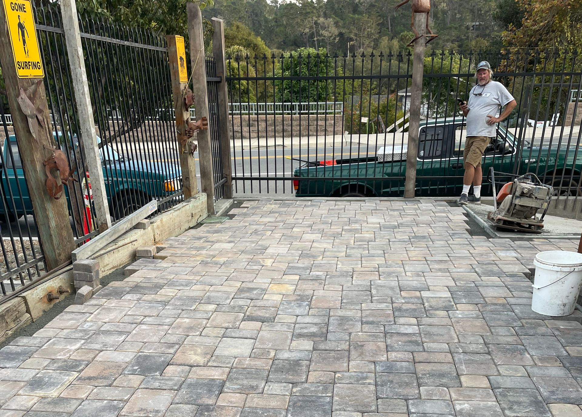 A worker stands on a newly laid gray stone paver patio next to a black metal fence with a park area in the background.