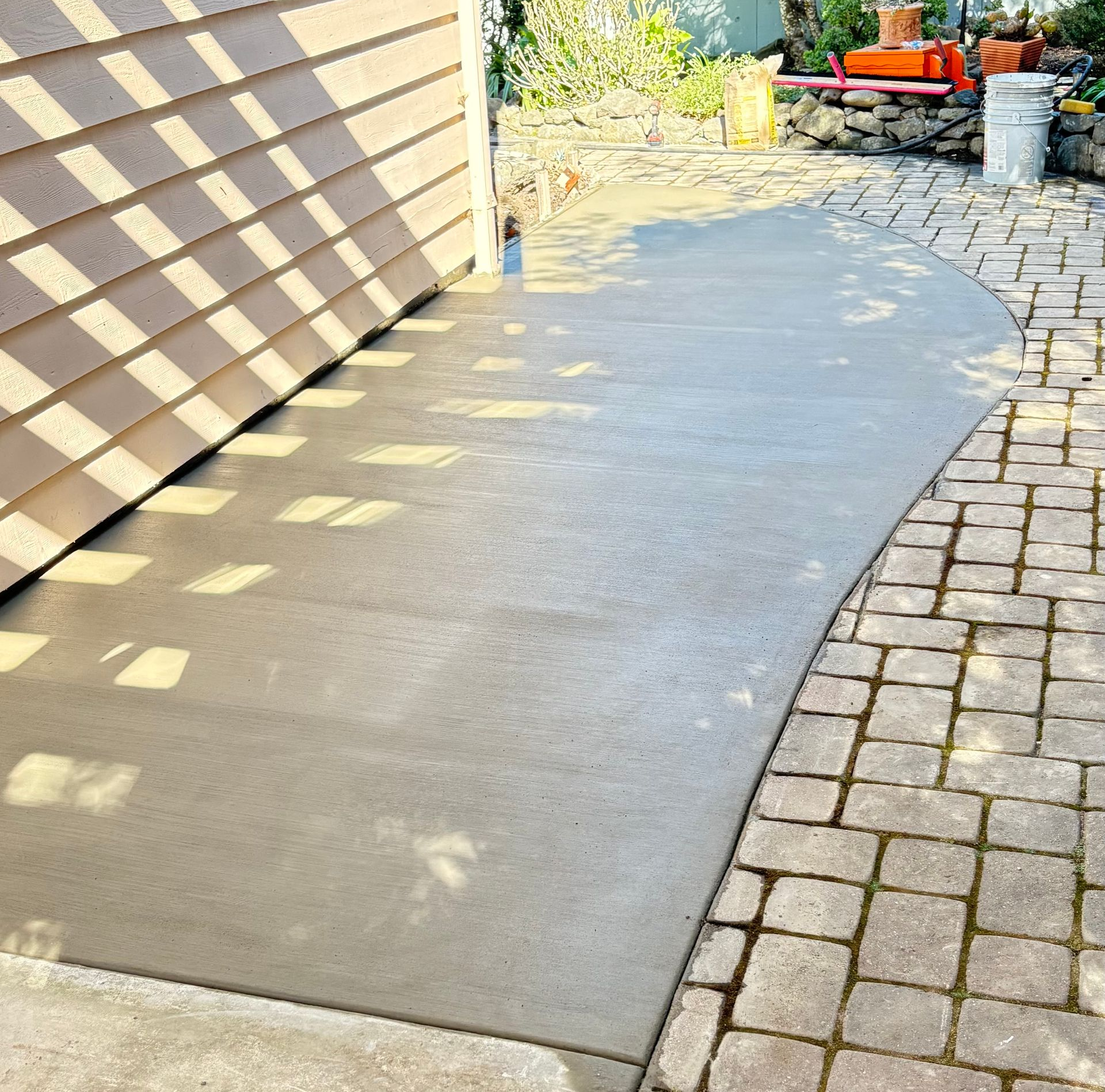 A freshly poured smooth concrete walkway curves beside a tan-sided wall and a stone paver patio.