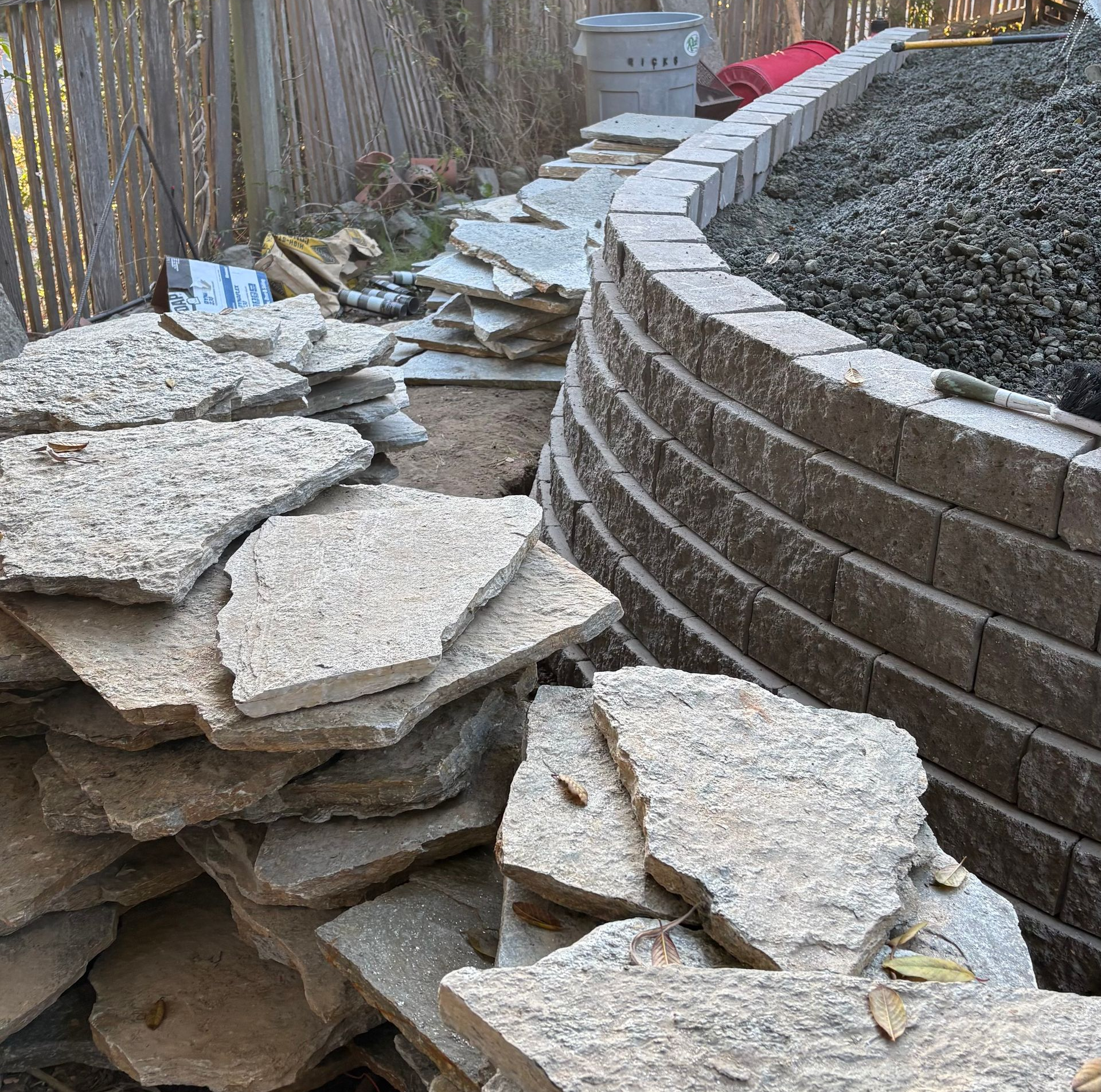 A stack of irregular, gray stone flagstones sits in front of a curved, tiered garden retaining wall made of tan bricks.