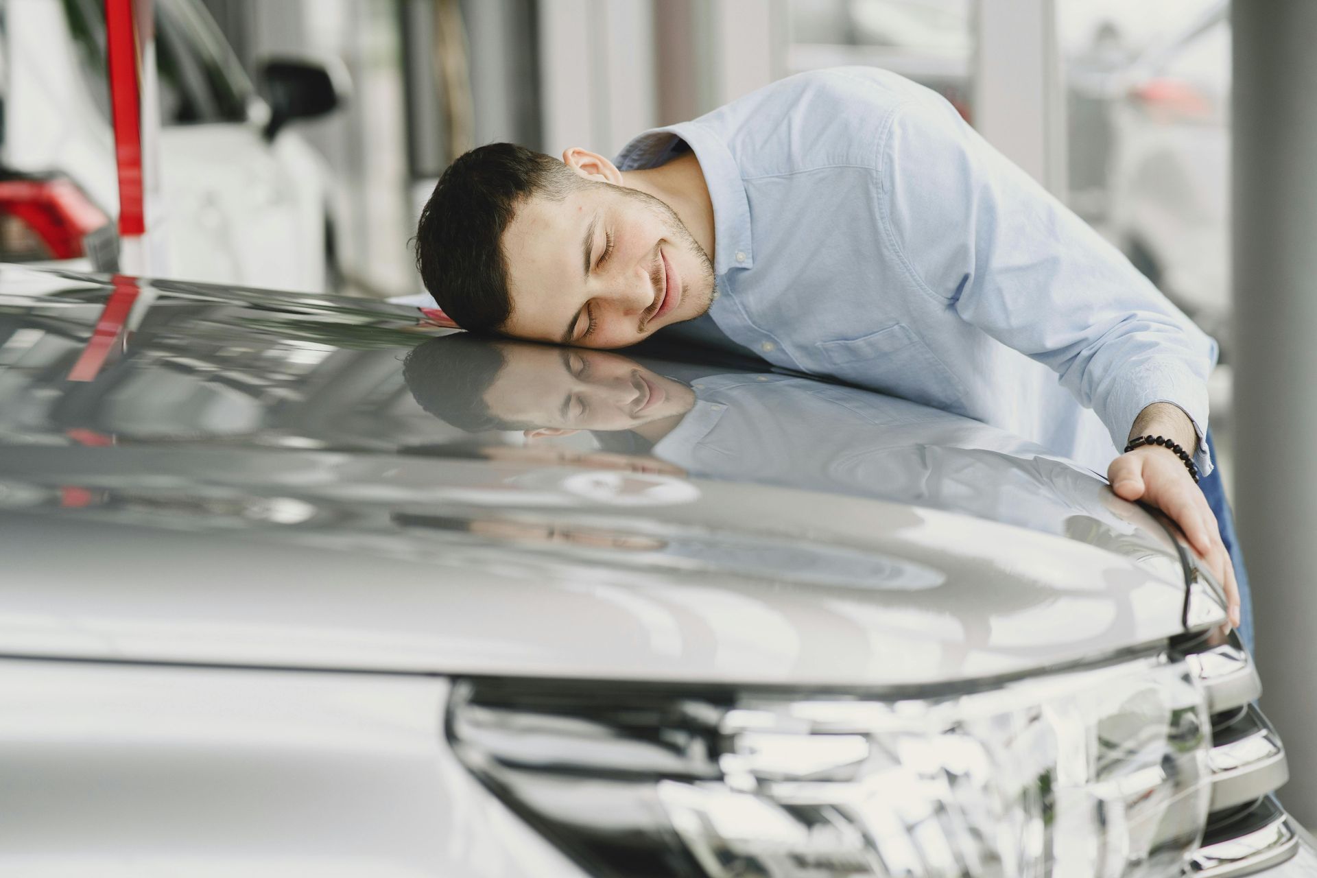 Man resting head on a car's hood in a car dealership, smiling.