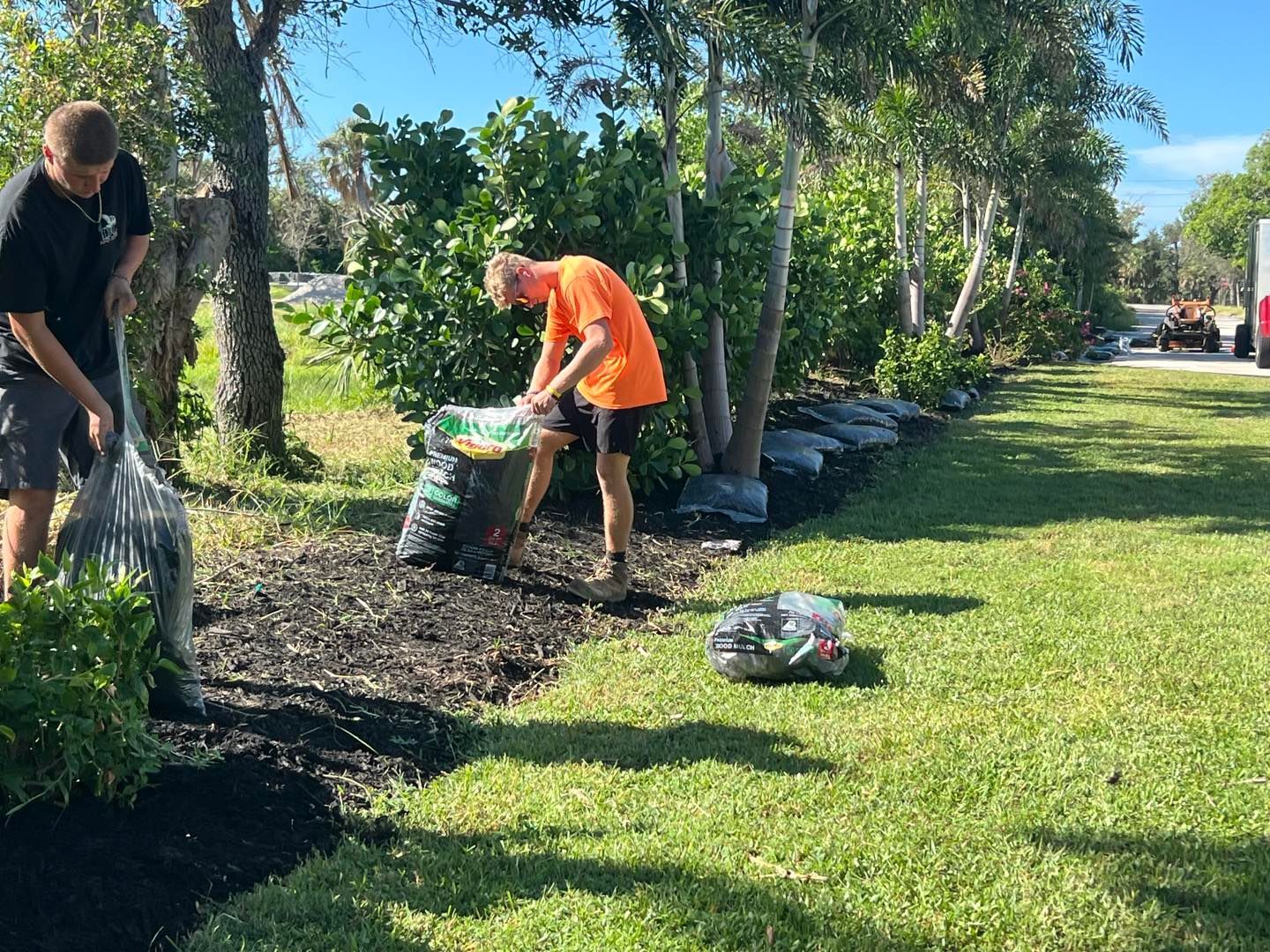 Two people bagging leaves beside a green lawn and garden bed on a sunny day