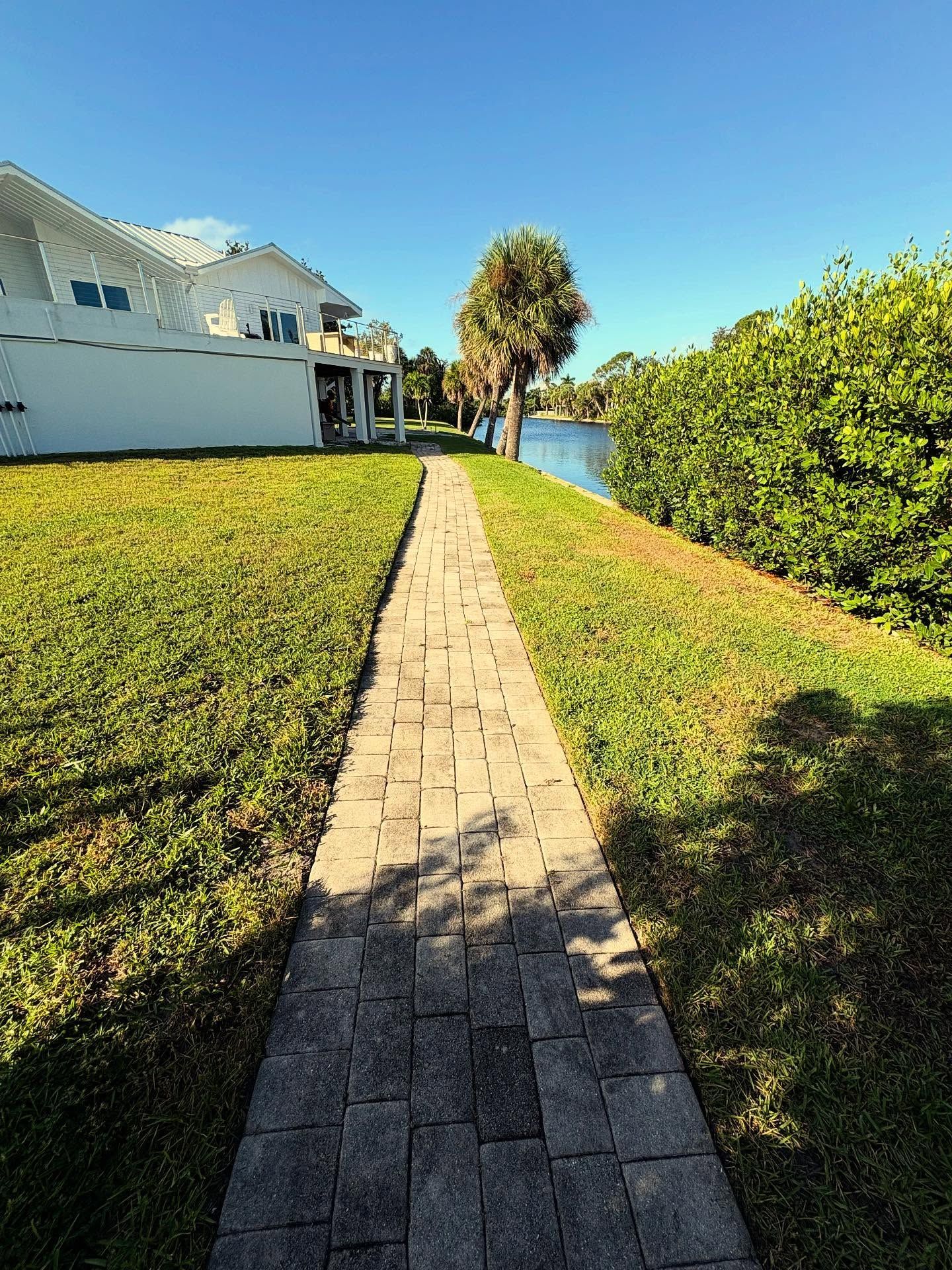 Brick walkway beside a white house, leading through green grass toward a lake under a bright blue sky