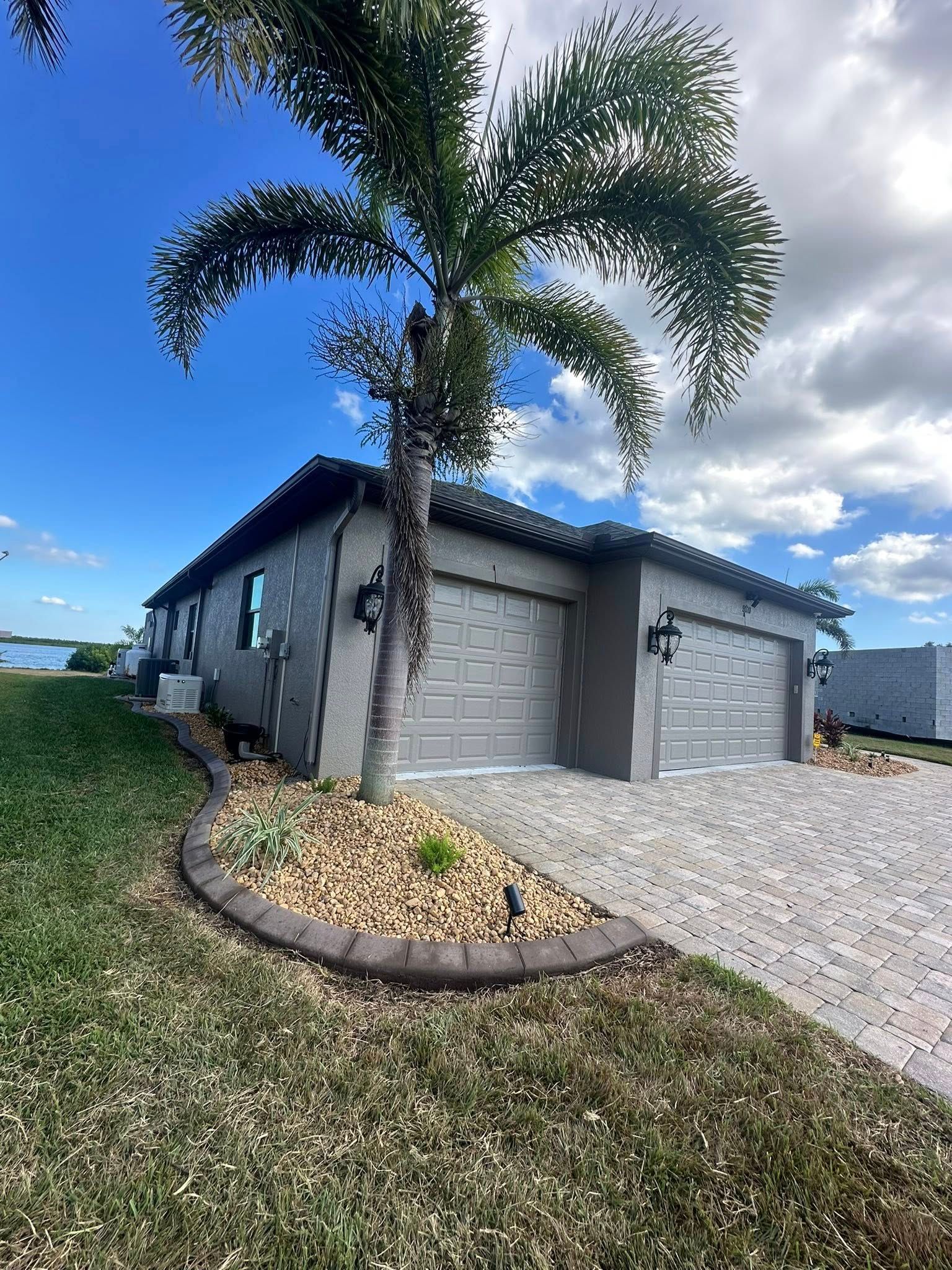Single-story gray house with palm tree, gravel driveway, and cloudy blue sky