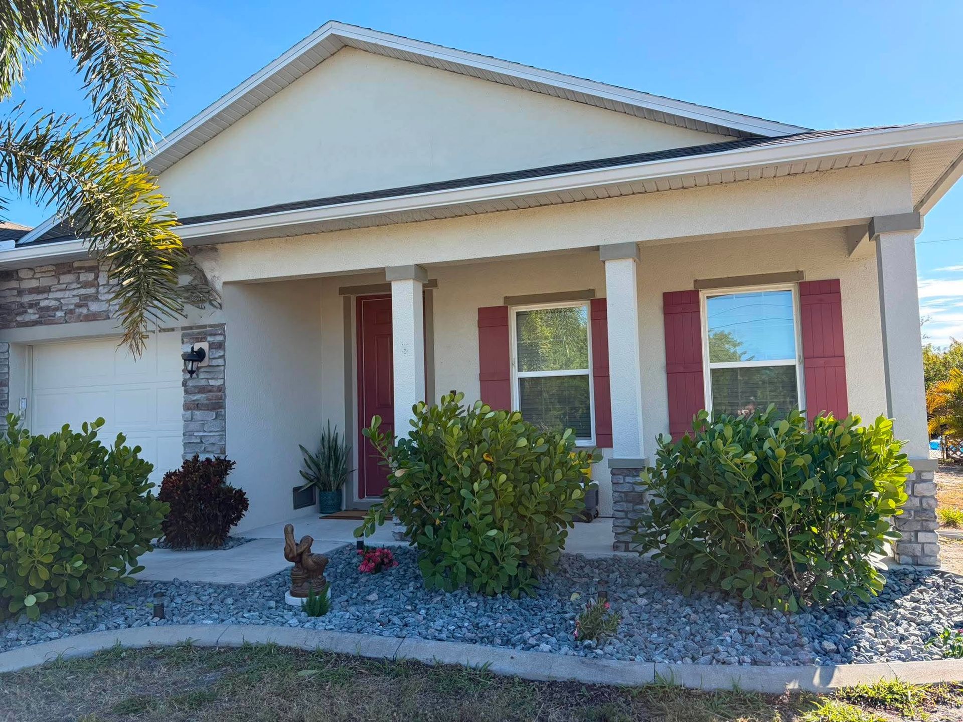 Single-story beige house with red shutters, front porch, and landscaped gravel yard