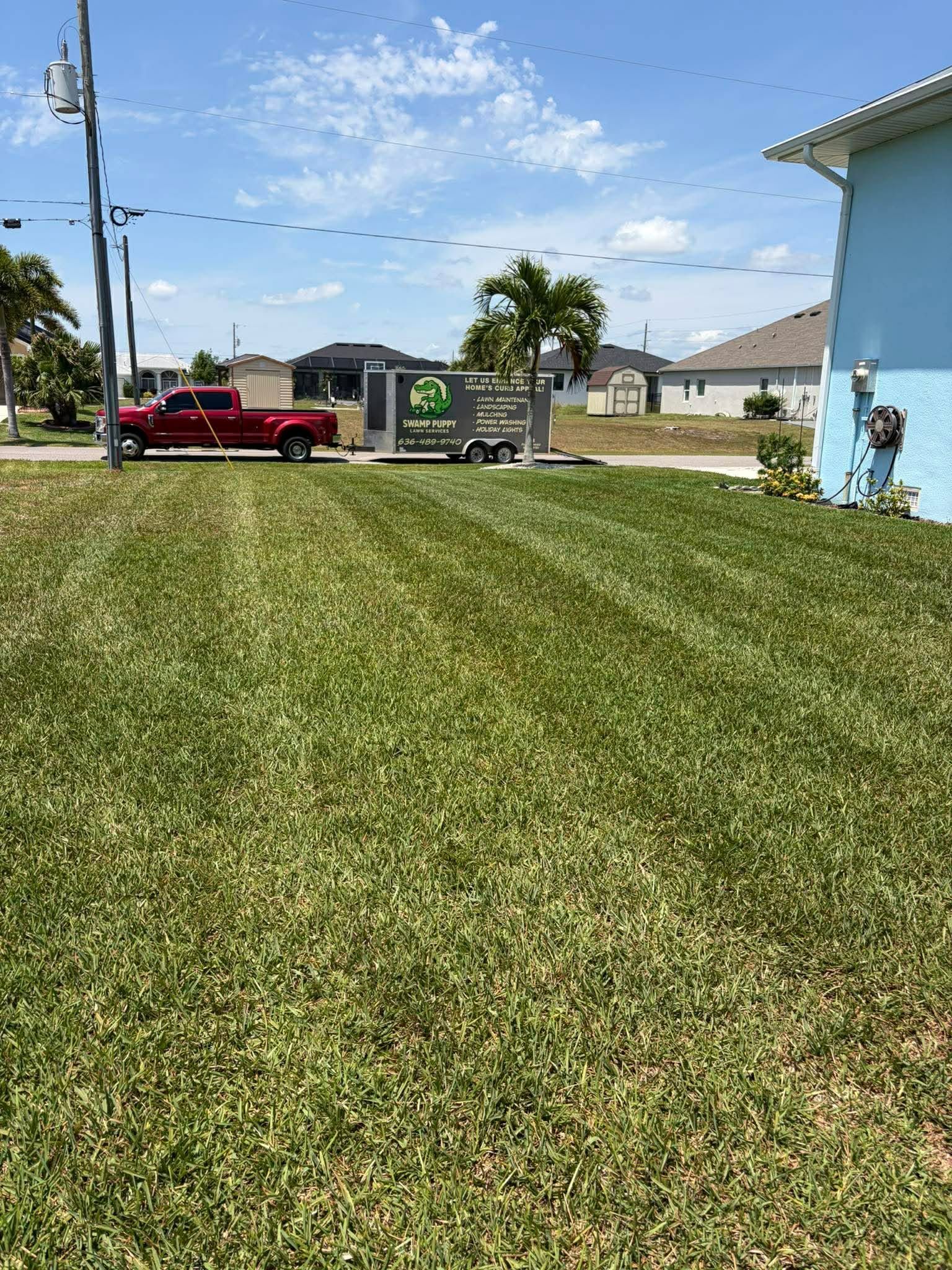 Wide grassy lawn with a red pickup truck and trailer near houses under a bright blue sky.