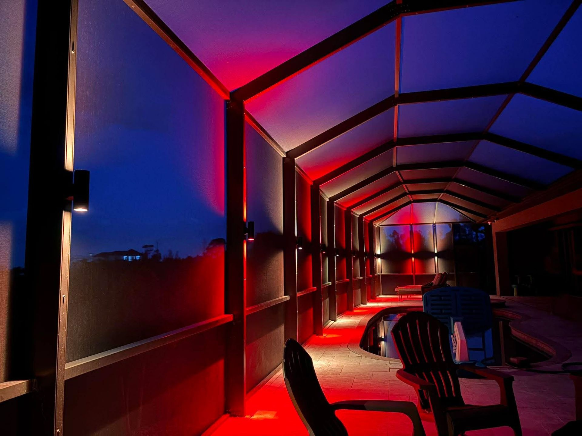 Red-lit covered walkway at dusk with chairs and a blue-purple sky beyond the railing