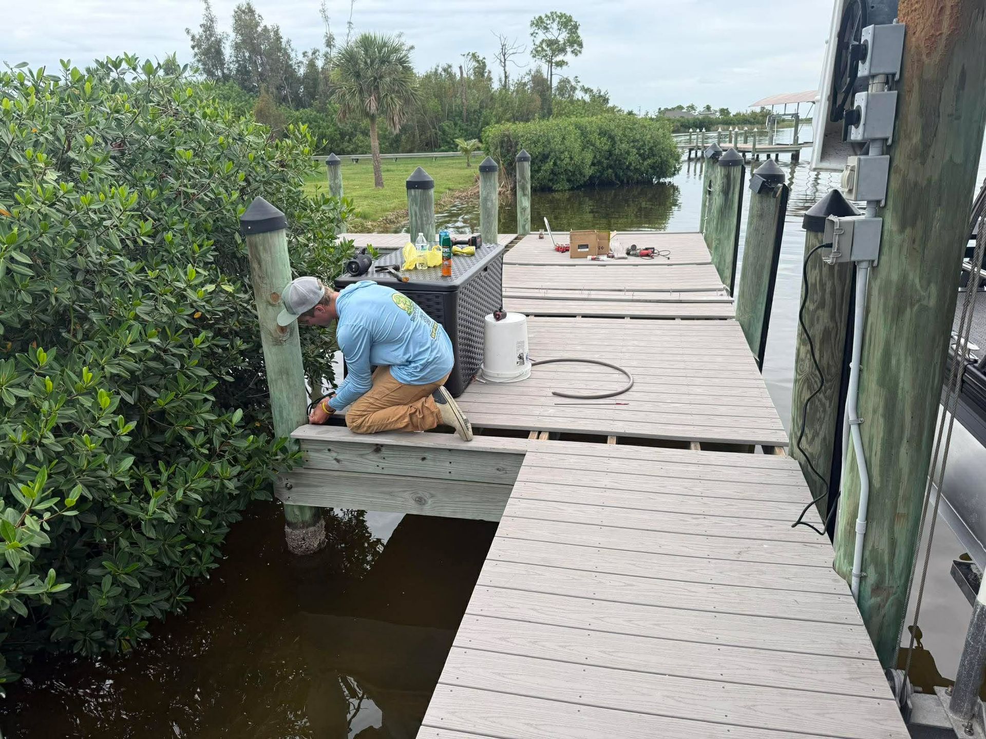 Worker on a dock repairing a wooden walkway beside water and mangrove trees.