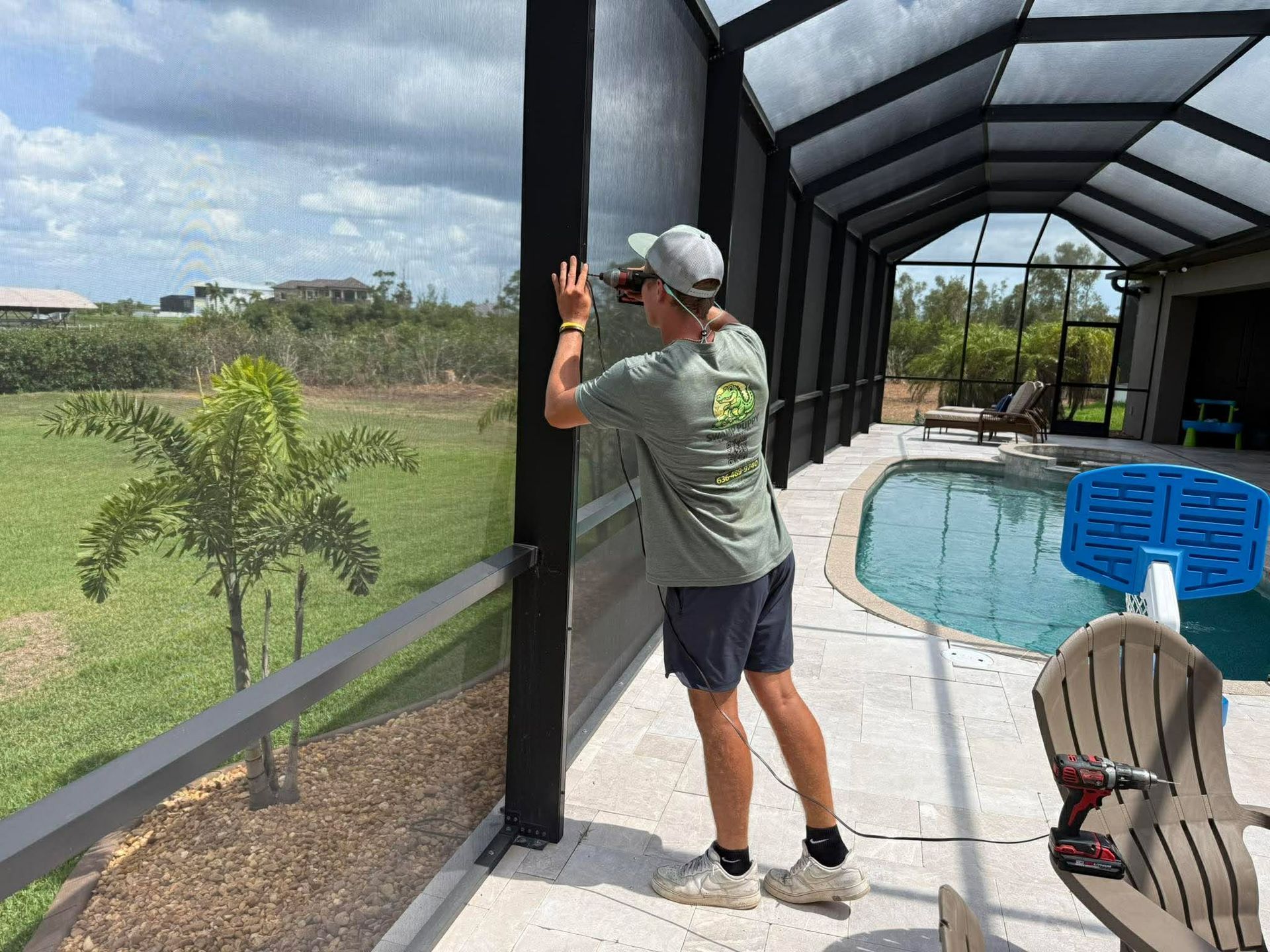 Person in gray shirt and cap stands by a screened patio overlooking a pool and green yard.