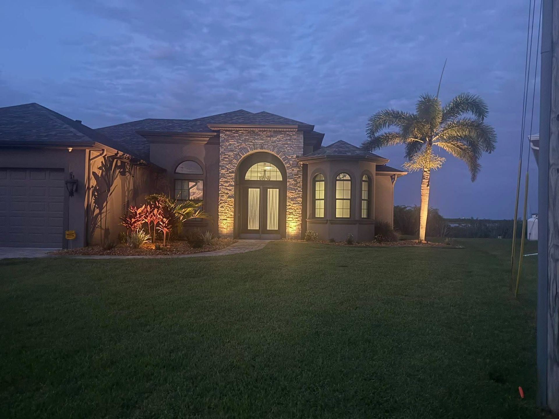 Luxury house at dusk with lit arched entryway, stone facade, palm tree, and wide green lawn
