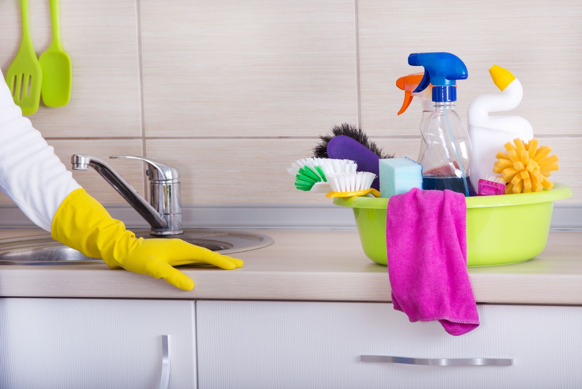 Gloved hand cleaning a kitchen counter beside a sink and a basket of colorful cleaning supplies