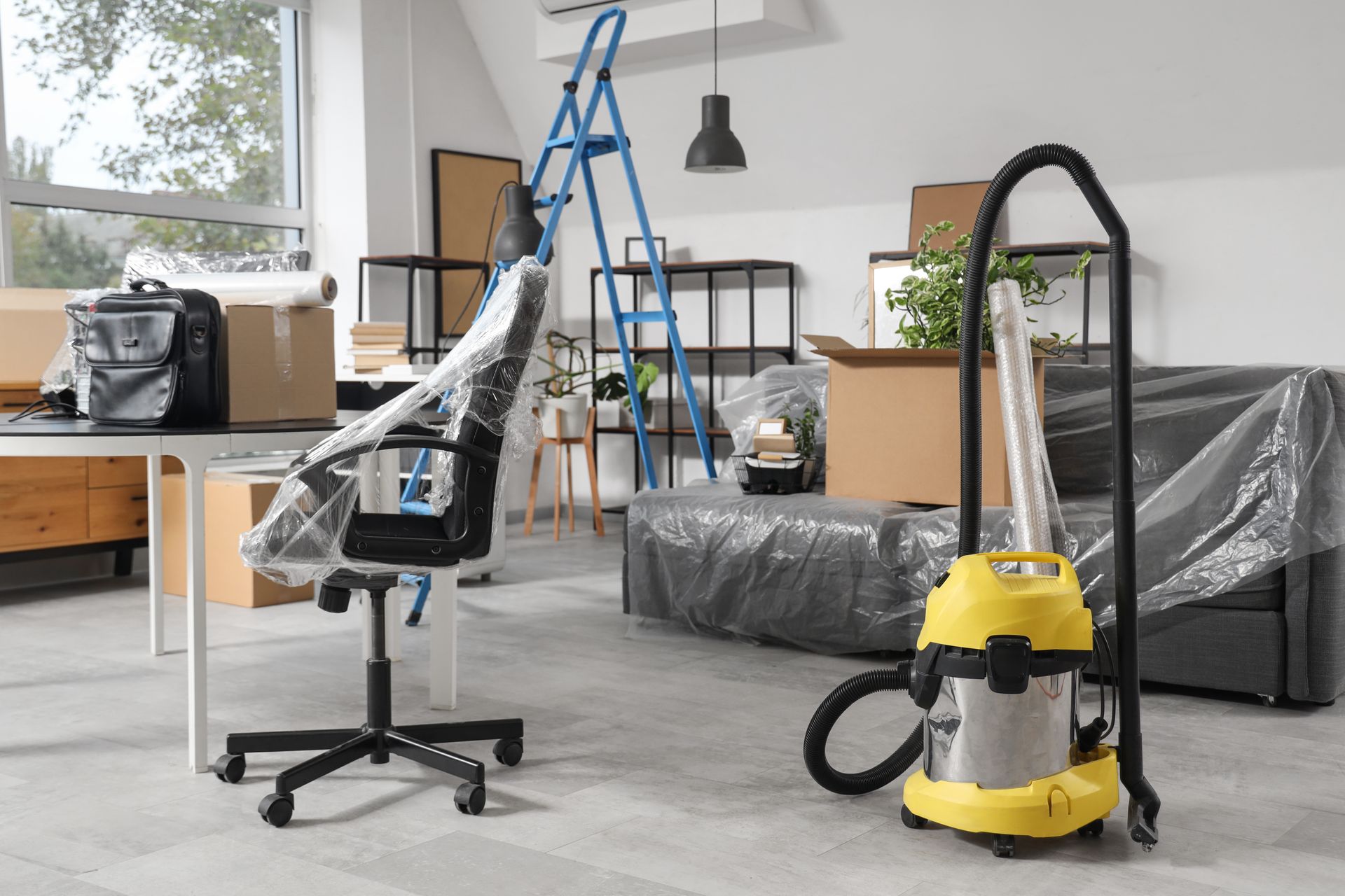 Home office under renovation with ladder, plastic-covered sofa, desk chair, and shop vacuum