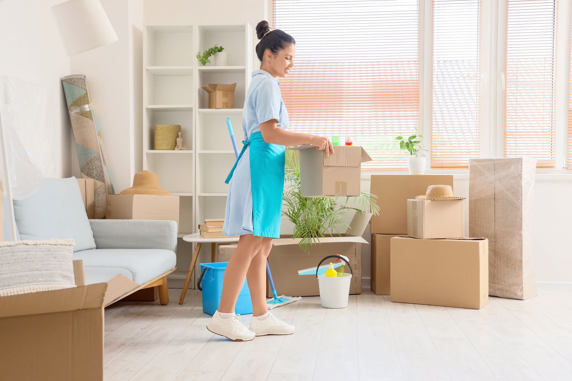 Person cleaning a bright living room with a spray bottle, surrounded by moving boxes and packing supplies