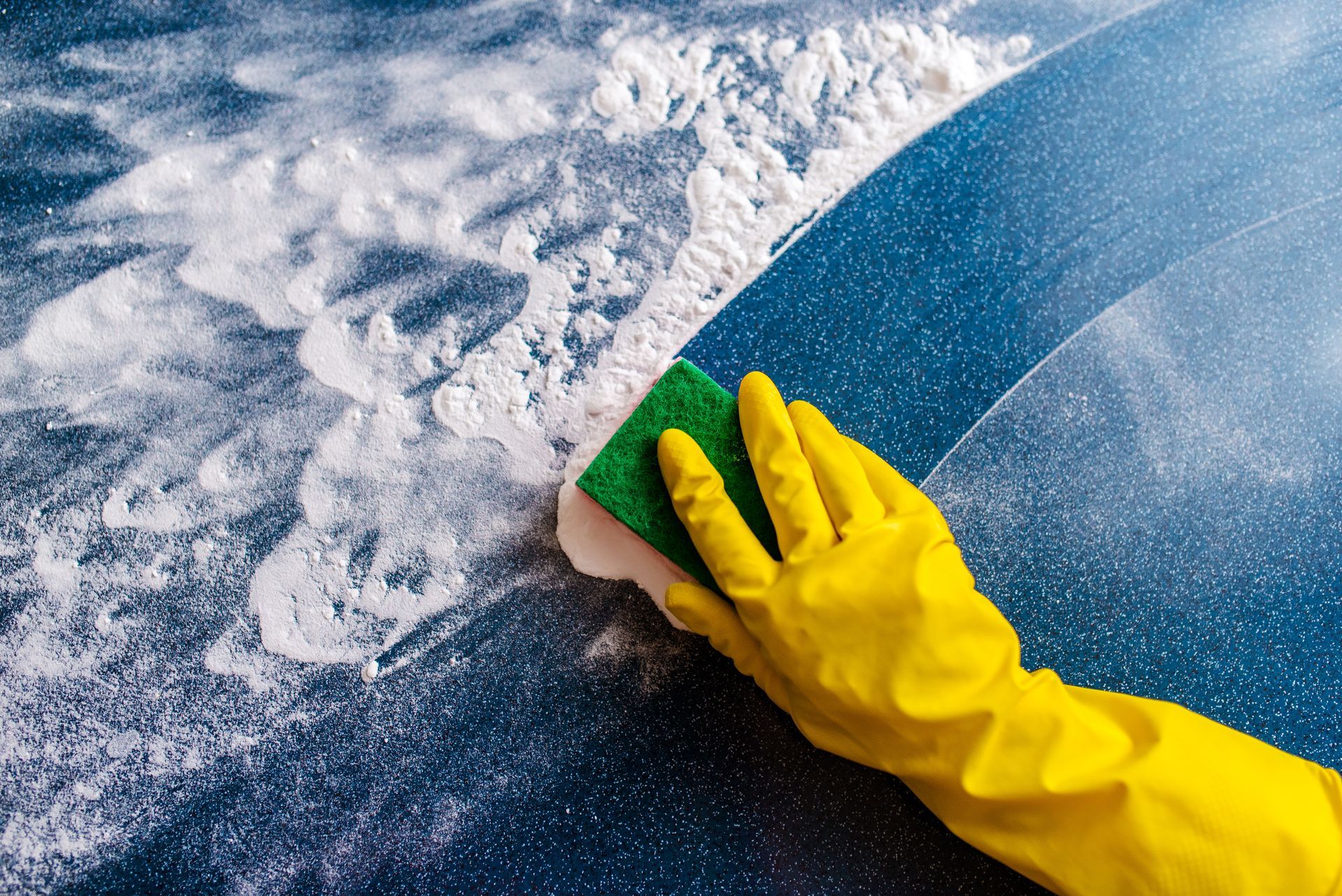 Yellow-gloved hand scrubbing a blue surface with a sponge and white foam