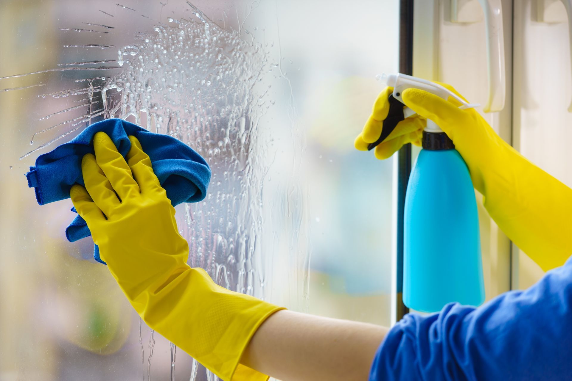 Yellow-gloved hands wiping a glass window with a blue cloth and spray bottle nearby